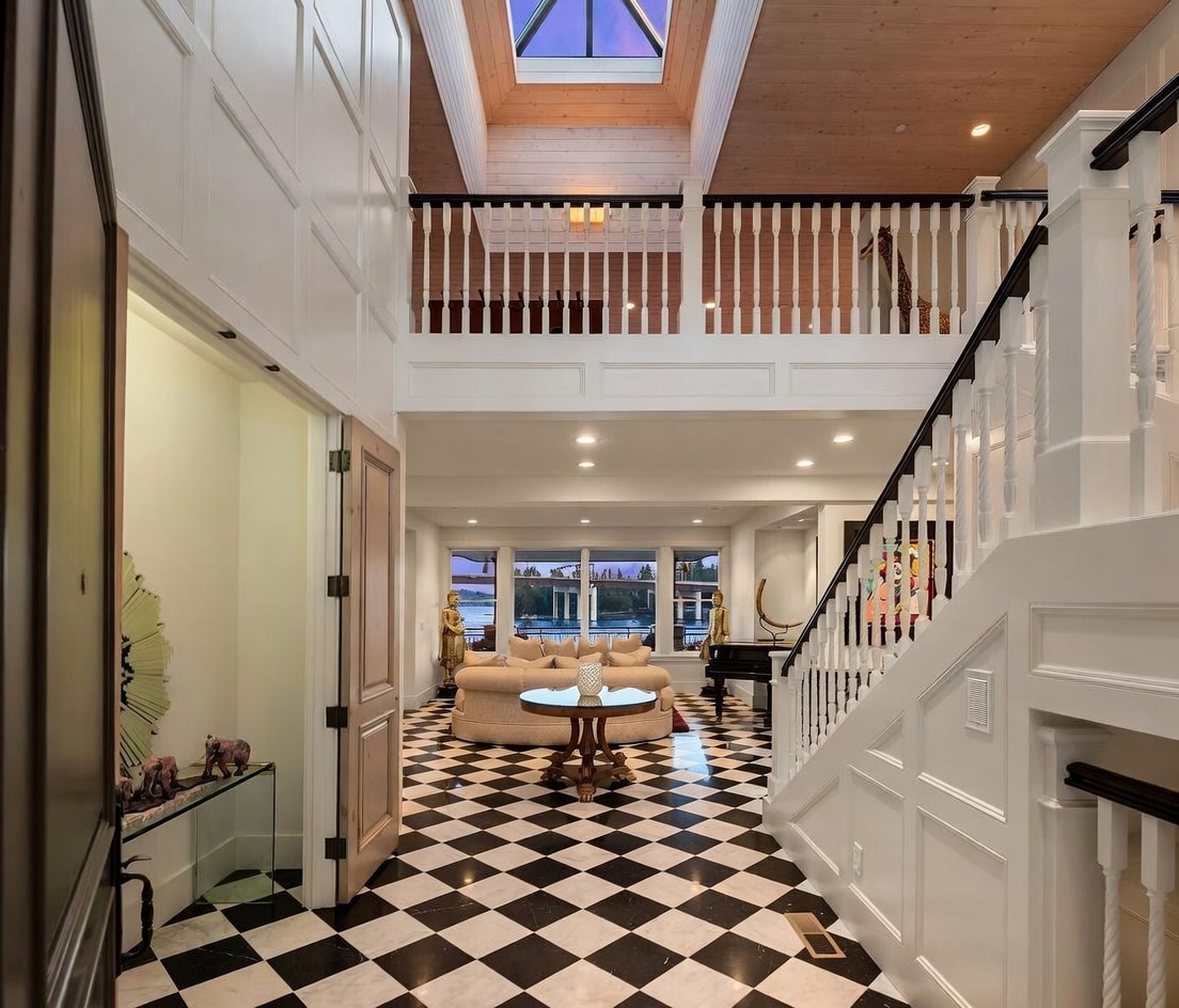 This grand entryway features a striking black-and-white checkered floor and a dramatic two-story ceiling with a central skylight. A white staircase with a dark handrail leads to an upper landing, while the open floor plan reveals a living area with large windows overlooking a waterfront view. The space is decorated with unique sculptural accents, creating an elegant and sophisticated atmosphere.