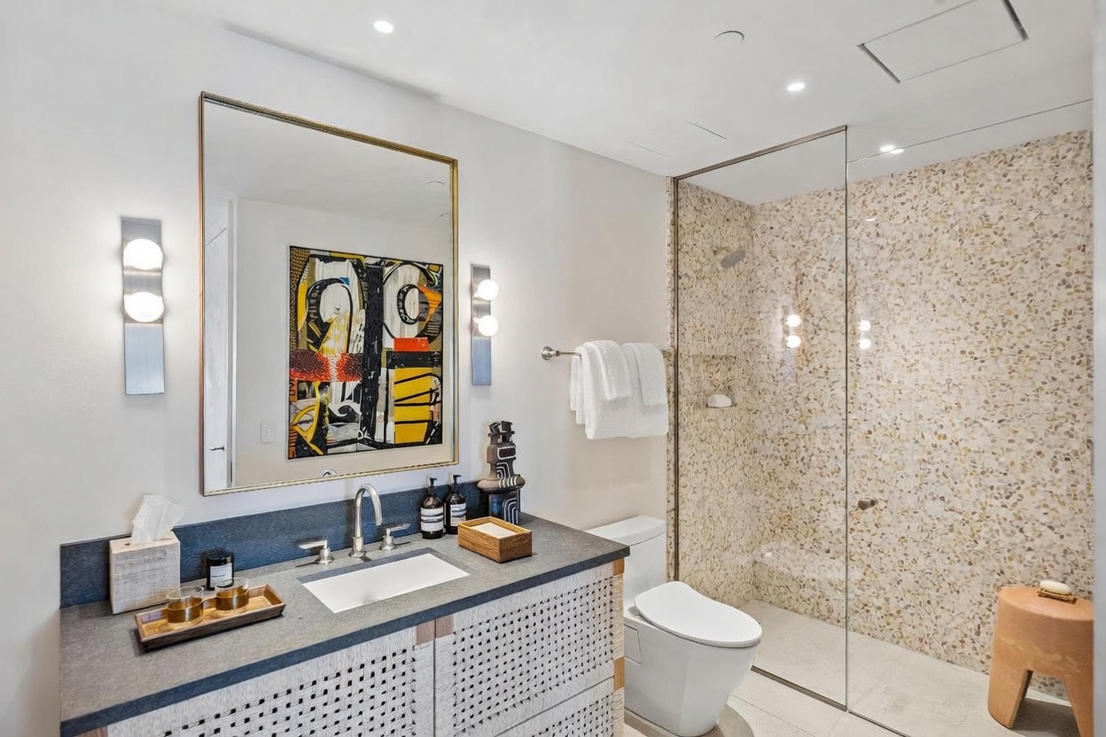 This modern bathroom features a vanity with a dark stone countertop and a textured, light-colored base, paired with a large rectangular mirror and contemporary wall sconces. To the right, a glass-enclosed walk-in shower showcases a distinctive, multi-colored speckled tile wall. The space is bright and well-lit, offering a clean, sophisticated aesthetic with a minimalist toilet and a small wooden stool in the shower area.