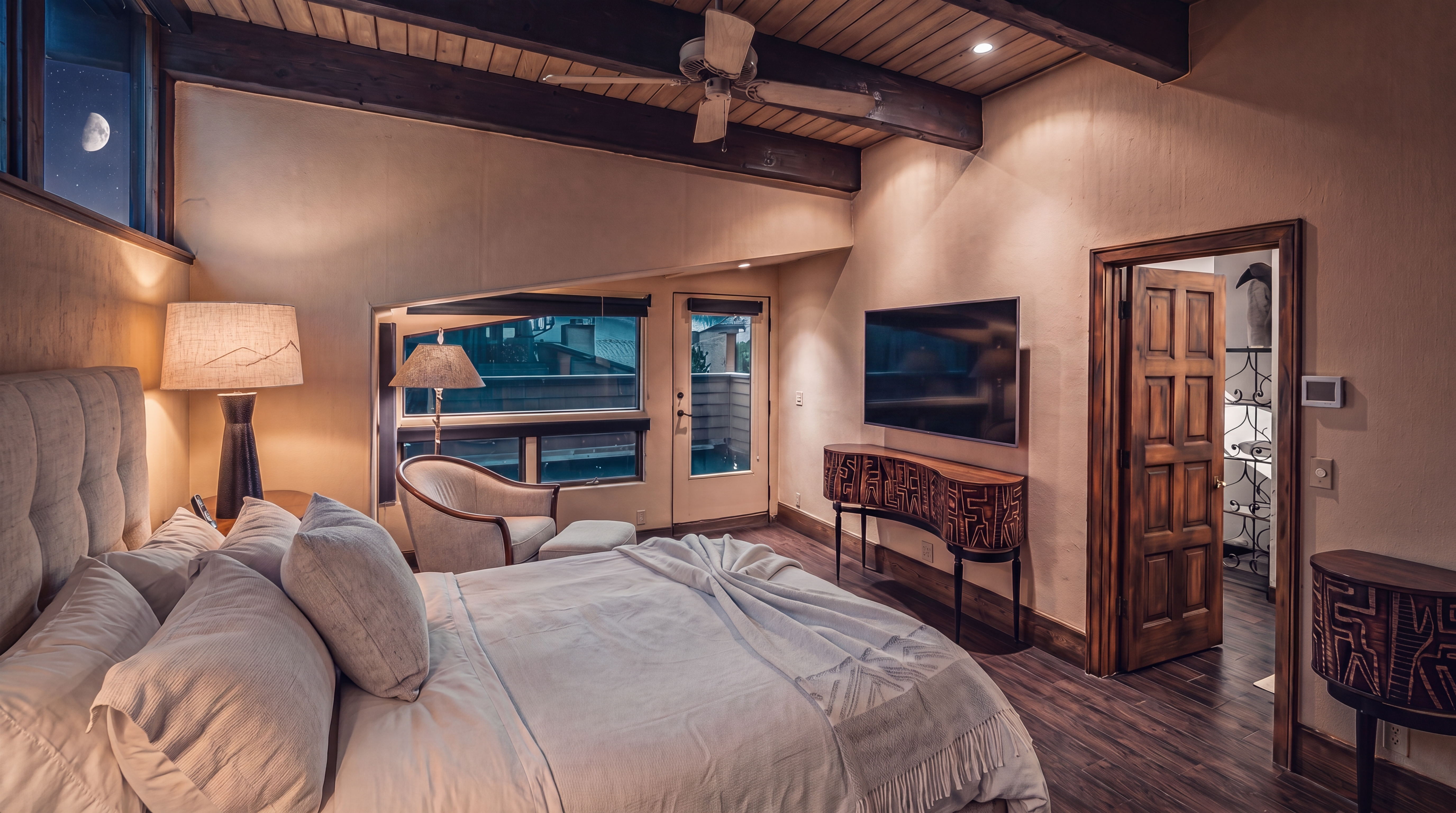 This primary bedroom features a vaulted ceiling with exposed dark wood beams and a central ceiling fan. The room is furnished with a large bed, a patterned console table beneath a wall-mounted television, and a cozy seating area by the windows. A wooden door leads to an adjacent bathroom, while the warm-toned walls and wood-look flooring create a comfortable, rustic atmosphere.
