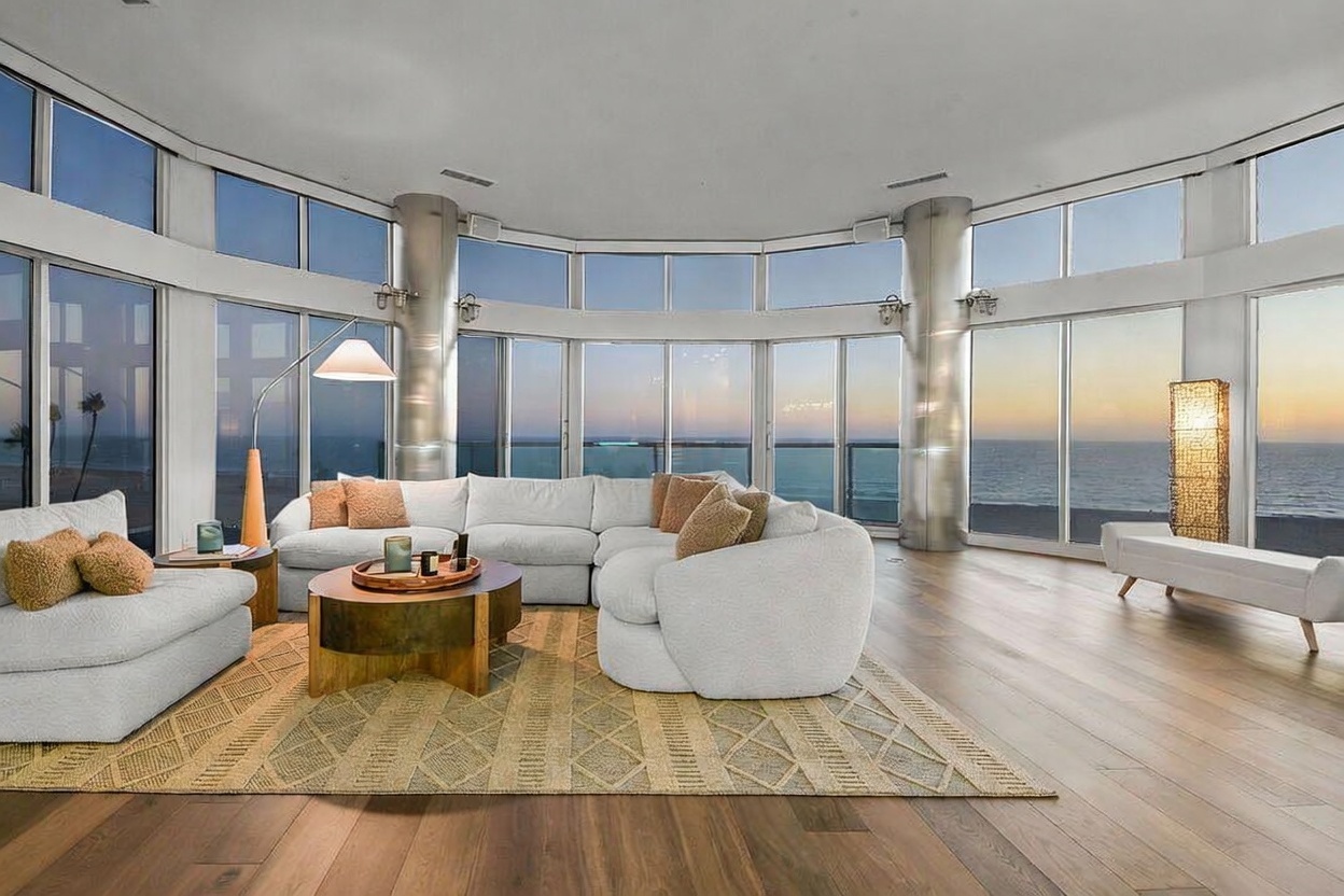 This expansive living room features a curved wall of floor-to-ceiling windows that provide panoramic ocean views. The space is anchored by a large, light-colored sectional sofa and a circular coffee table, all set upon a textured area rug over light wood flooring. Two metallic structural columns frame the view, contributing to a modern, open-concept aesthetic that emphasizes the connection between the interior living space and the coastal horizon.