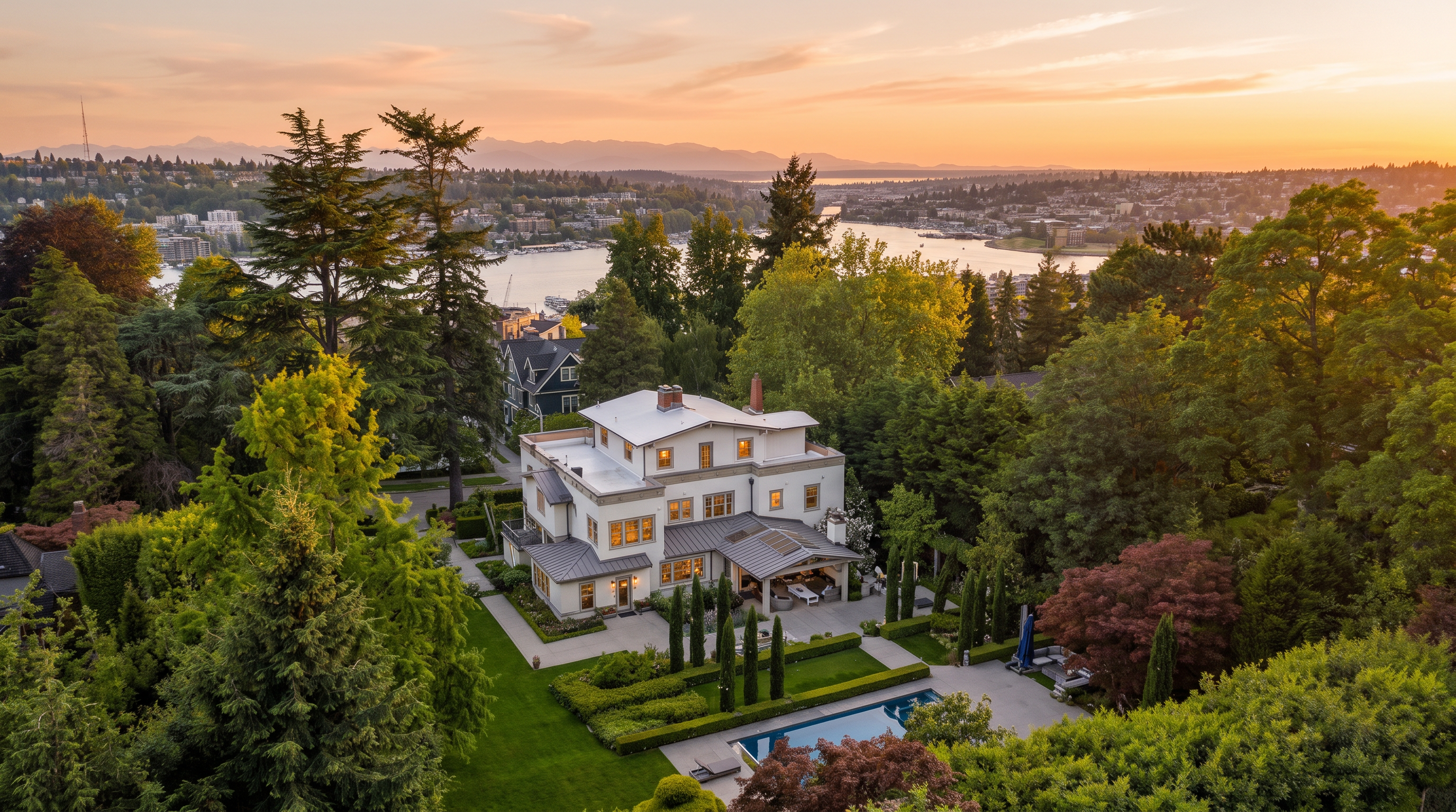 This stunning high-angle aerial photograph showcases a luxury estate overlooking a scenic lake and mountains during a vibrant sunset. The property features a multi-story home with classic architecture, expansive manicured lawns, formal landscaping, and a pristine swimming pool. The lush surroundings and panoramic water view create an exceptionally serene and high-end residential atmosphere.