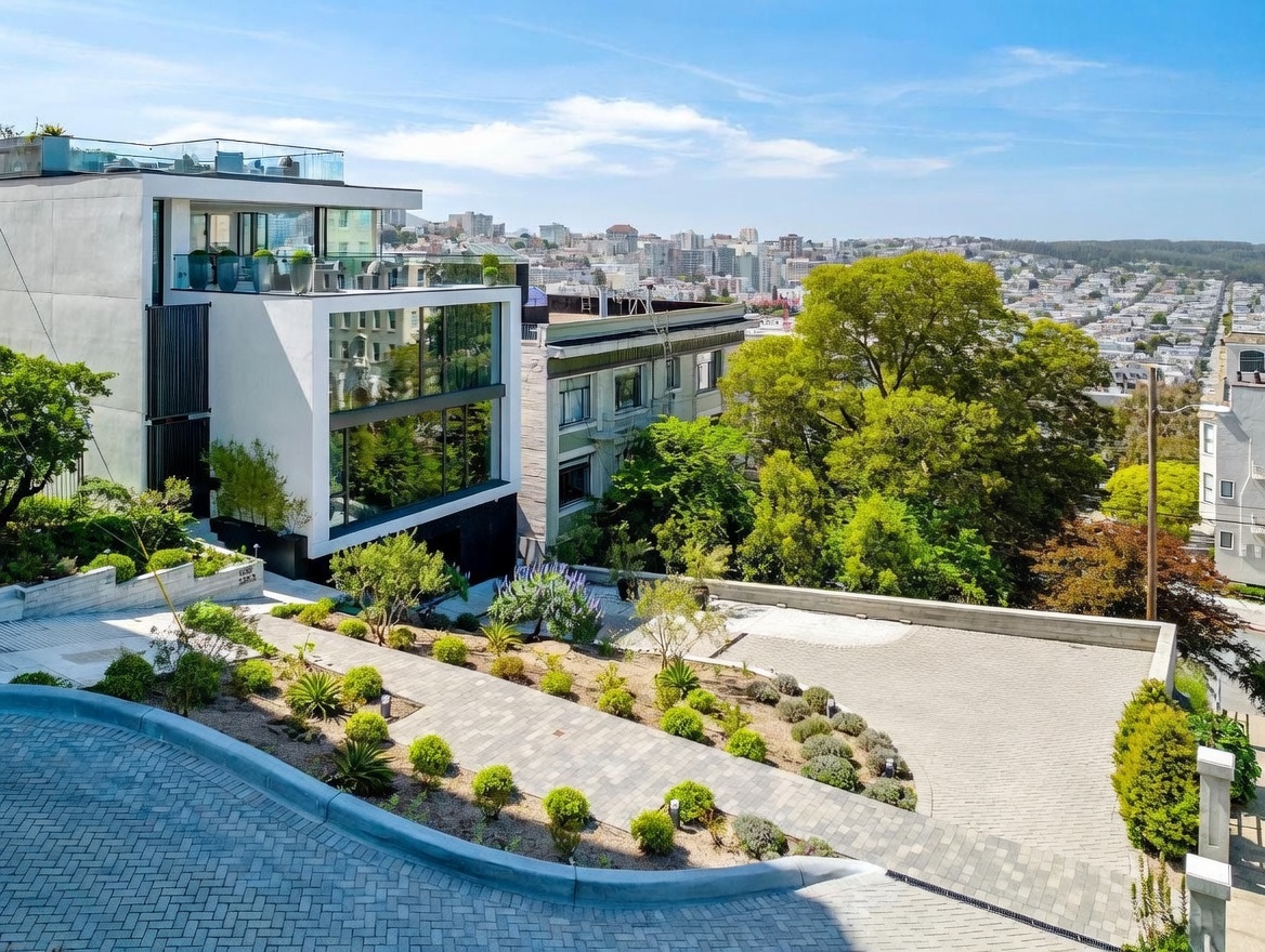 This modern, multi-level residence features a striking architectural design with clean white lines, dark accents, and expansive glass walls that offer panoramic city views. The property is situated on a sloped lot, incorporating a tiered landscape with paved walkways, manicured shrubs, and a spacious driveway area. The perspective is elevated, capturing the home's integration into the hillside and its prominent position overlooking the urban skyline.