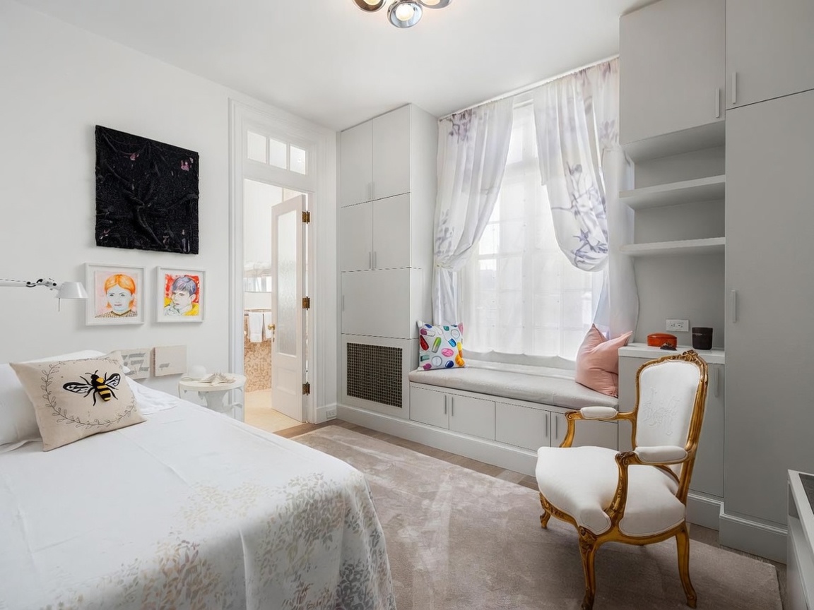 This bright and airy bedroom features a plush bed adorned with a bee-themed throw pillow and a classic gold-leaf accent chair. A built-in window seat with integrated storage cabinetry provides a cozy nook, framed by sheer curtains that allow soft natural light to fill the room. The space is decorated with eclectic wall art and leads directly into an adjacent bathroom through a white paneled door.