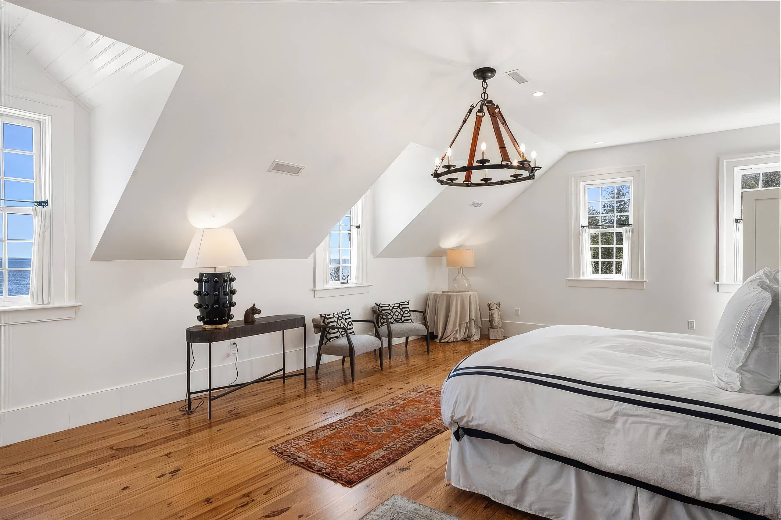 This bright and airy primary bedroom features a vaulted ceiling with dormer windows that offer scenic water views. The room is furnished with a large bed, a sleek console table with a decorative lamp, and two armchairs arranged near a small round table. A warm-toned patterned rug anchors the space, which is finished with light wood flooring and a rustic-style chandelier.