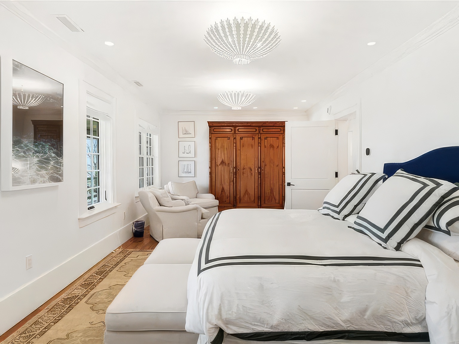 This bright and airy primary bedroom features a large bed with a navy blue headboard and white bedding accented by dark stripes. A pair of comfortable armchairs sits near a large wooden wardrobe, while a patterned rug adds texture to the hardwood floors. The room is illuminated by two distinctive, textured pendant lights, creating a sophisticated and serene atmosphere.