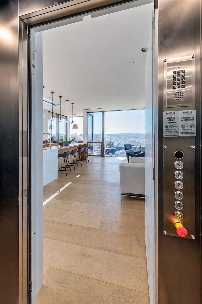 This image is taken from inside a private elevator looking out into a modern, open-concept living space. The foreground features the metallic elevator control panel with a button array, while the background reveals a bright kitchen with a long island, bar seating, and expansive windows showcasing a city view. The interior design is characterized by light wood flooring and a clean, minimalist aesthetic that seamlessly connects the indoor living area with the urban landscape beyond.