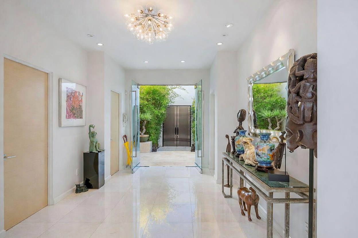 This elegant entryway features polished light-colored tile flooring and a bright, airy atmosphere leading toward an open glass door that reveals a lush outdoor garden. The space is decorated with artistic sculptures and a long console table topped with decorative vases, all reflected in a large, ornate mirror. A modern, multi-bulb chandelier hangs from the ceiling, casting a warm glow over the sophisticated, gallery-like foyer.