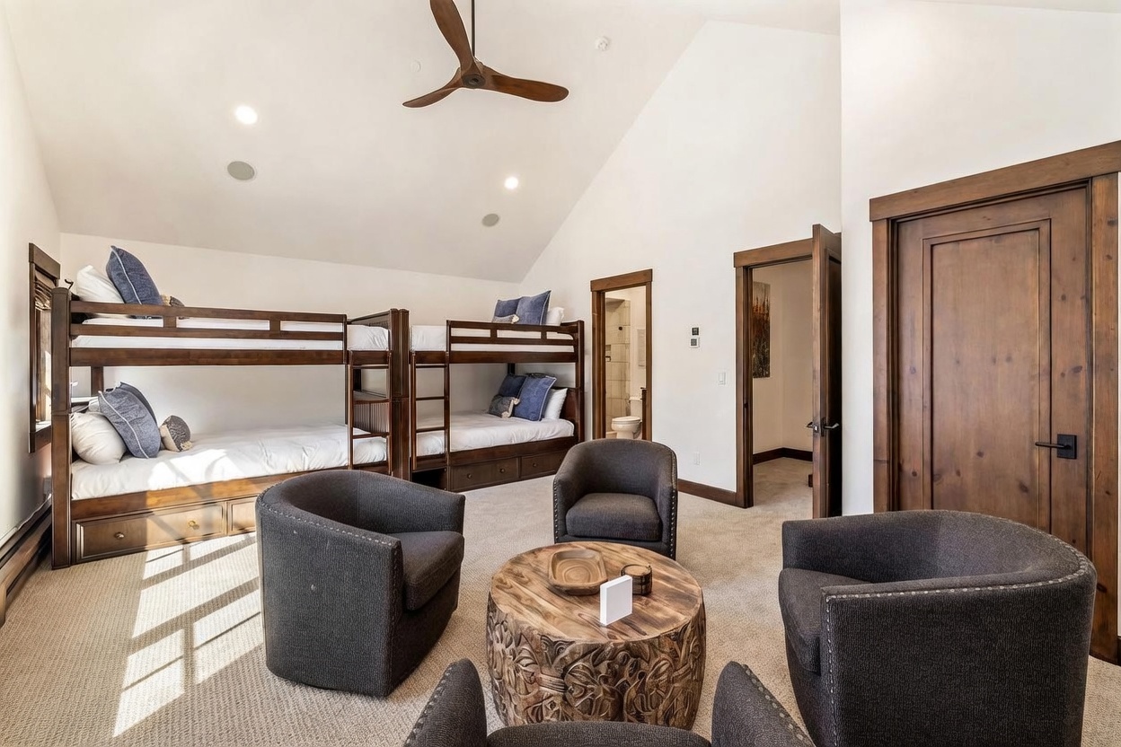 This spacious bedroom features a rustic-modern aesthetic with vaulted ceilings and a large bunk bed unit that sleeps four. A cozy seating area is arranged in the center of the room, consisting of four dark upholstered armchairs surrounding a carved wooden coffee table. The room is finished with neutral carpeting, warm wood trim, and provides direct access to an en-suite bathroom and a secondary doorway.