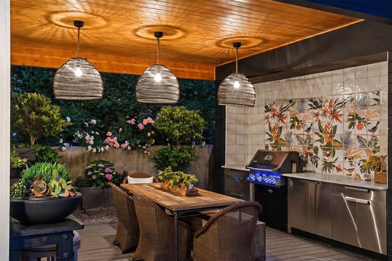 This inviting outdoor dining and kitchen area features a covered deck with a warm wood-paneled ceiling and three decorative wire pendant lights. The space includes a stainless steel grill built into a counter with a vibrant, patterned tile backsplash, alongside a rustic wooden dining table and wicker chairs. Lush greenery and blooming flowers in the background create a serene, private atmosphere perfect for alfresco entertaining.