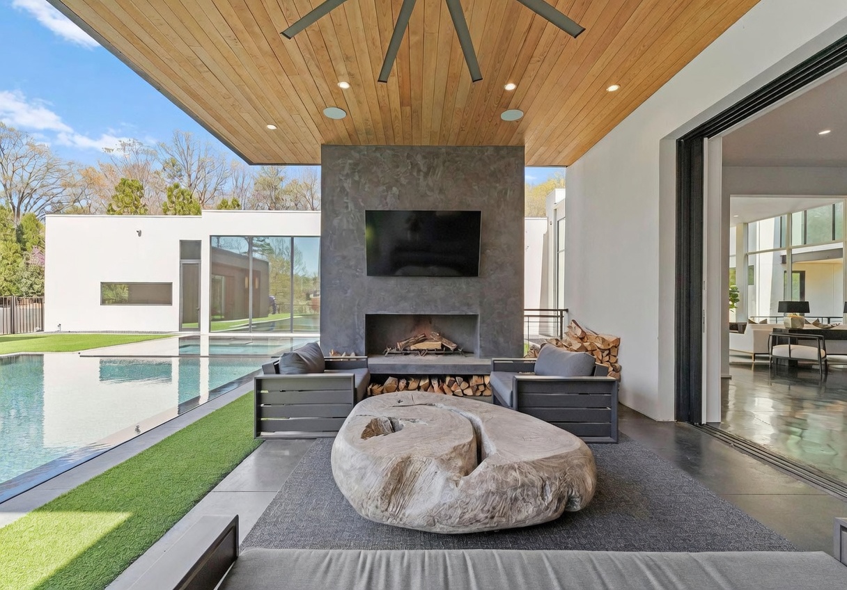 This luxurious covered outdoor living space features a prominent dark stone chimney breast with a built-in fireplace and a mounted television. The area is furnished with modern gray seating and a unique, large organic-shaped wooden coffee table, all set beneath a warm-toned wood-paneled ceiling. The space seamlessly transitions to a pristine swimming pool and manicured lawn, framed by large glass sliding doors that open into the interior living area.