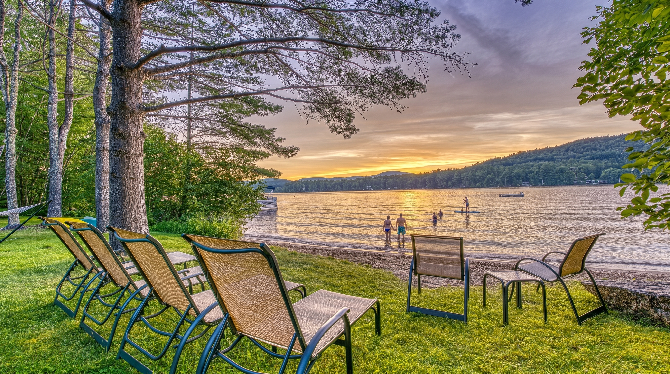 This serene waterfront scene captures a grassy lawn furnished with several outdoor lounge chairs facing a calm lake at sunset. Tall, mature trees frame the left side of the composition, while the golden light of the setting sun reflects off the water's surface. The perspective is low and wide, emphasizing the peaceful connection between the private yard and the natural shoreline.