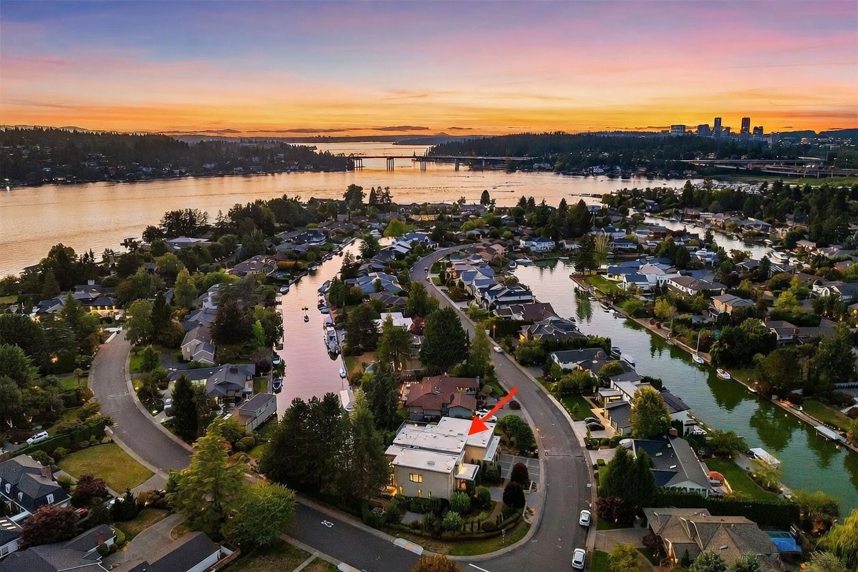 This high-angle aerial view captures a residential property nestled within a waterfront neighborhood at sunset. The scene features a winding canal, lush landscaping, and a distant city skyline under a vibrant orange and purple sky. A red arrow highlights the specific property, which is situated on a corner lot near the water's edge.