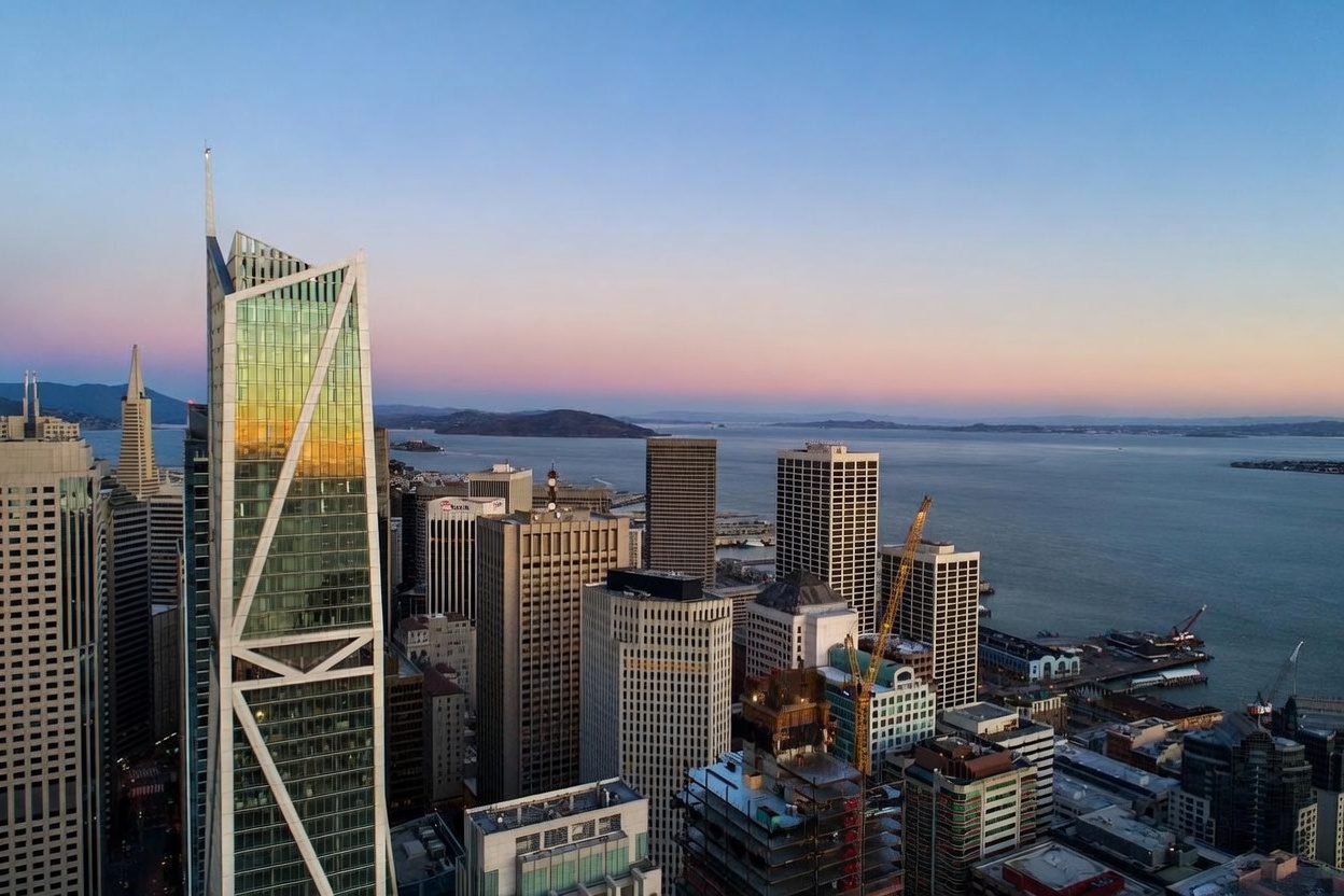This high-altitude aerial view captures the dense urban skyline of San Francisco at dusk, featuring the prominent, angular glass facade of a modern skyscraper in the foreground. The composition highlights the contrast between the reflective, geometric architecture of the city's high-rises and the expansive, calm waters of the bay stretching toward the horizon. The soft, gradient colors of the sunset sky provide a serene backdrop to the bustling metropolitan landscape.