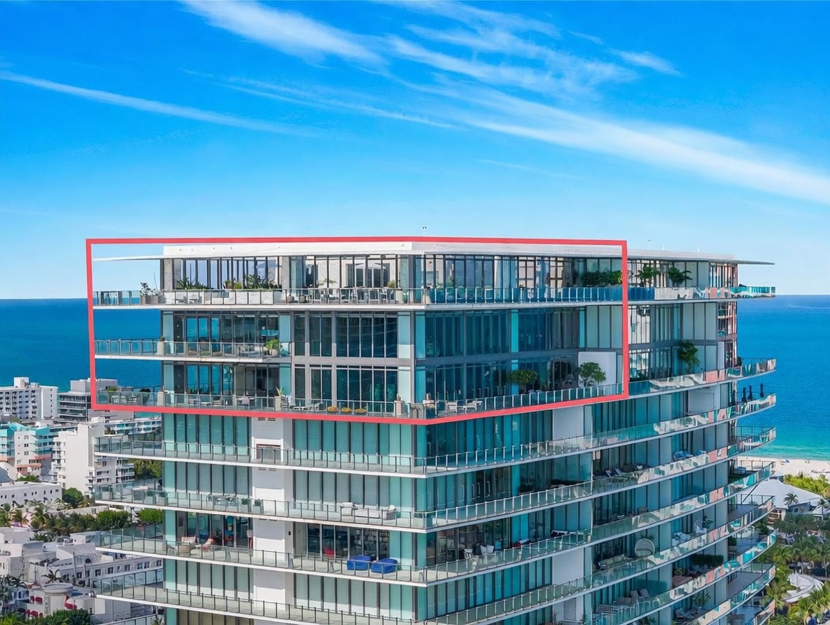 This aerial perspective captures the upper levels of a modern, high-rise luxury condominium building situated directly along the coastline. The structure features extensive glass facades and expansive balconies, with a red-outlined section highlighting the top-floor penthouse units that offer panoramic ocean views. The bright blue sky and turquoise water create a serene, upscale atmosphere typical of a premier beachfront property.