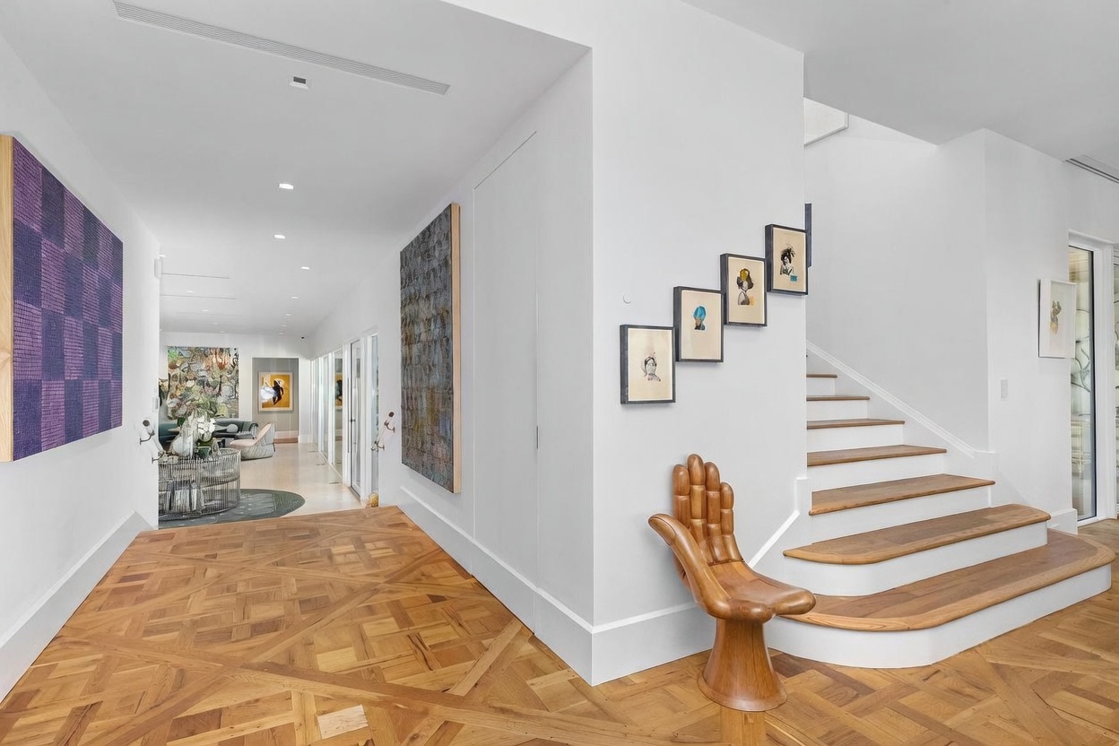 This elegant interior space features a wide hallway with intricate patterned wood flooring that leads toward a bright, open living area in the distance. To the right, a staircase with light wood treads ascends, accented by a unique hand-shaped wooden chair and a series of framed artworks on the wall. The aesthetic is sophisticated and gallery-like, characterized by clean white walls, recessed lighting, and a seamless transition between the foyer and the adjacent living spaces.