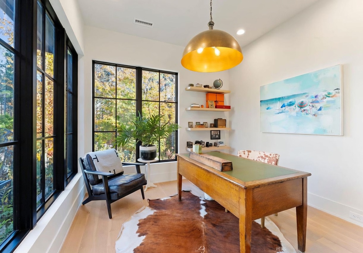This bright and airy home office features large, black-framed windows that offer a scenic view of the outdoors. The room is anchored by a vintage-style wooden desk with a green top, complemented by a cowhide rug and a dark leather armchair. Floating shelves provide organized storage for decor, while a large, gold-toned pendant light serves as a striking focal point above the workspace.