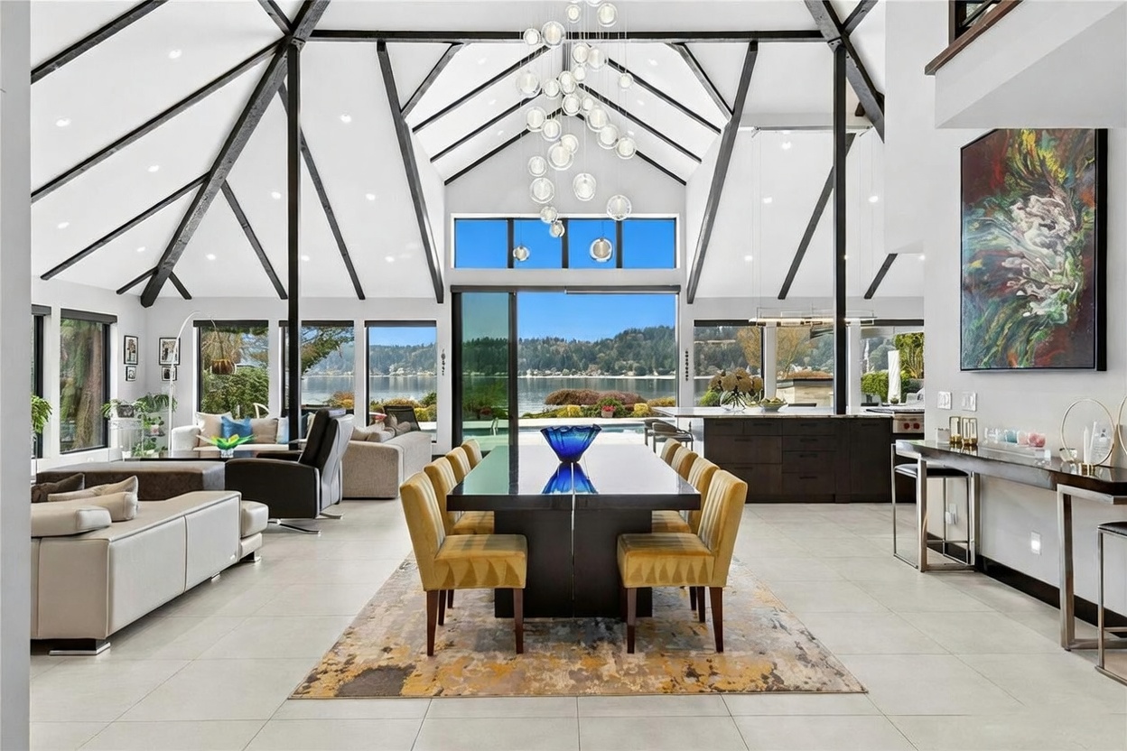 This expansive, open-concept dining area features a dramatic vaulted ceiling with exposed black beams and a striking modern chandelier. The room seamlessly transitions into a living space and kitchen, all centered around a large dining table and floor-to-ceiling windows that frame a stunning lake view. The aesthetic is sophisticated and contemporary, emphasizing natural light and a strong connection to the outdoor scenery.