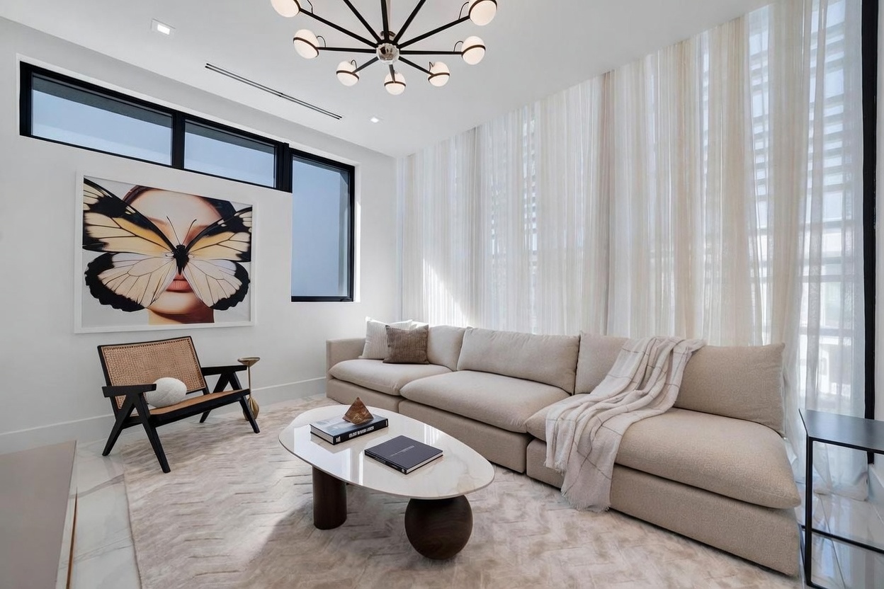 This bright and modern living room features a large, neutral-toned sectional sofa paired with a unique oval coffee table on a textured area rug. A stylish accent chair with cane detailing sits near a striking piece of butterfly-themed wall art, while sheer curtains drape over tall windows that allow for ample natural light. The space is anchored by a contemporary multi-arm chandelier, creating a sophisticated and airy atmosphere.