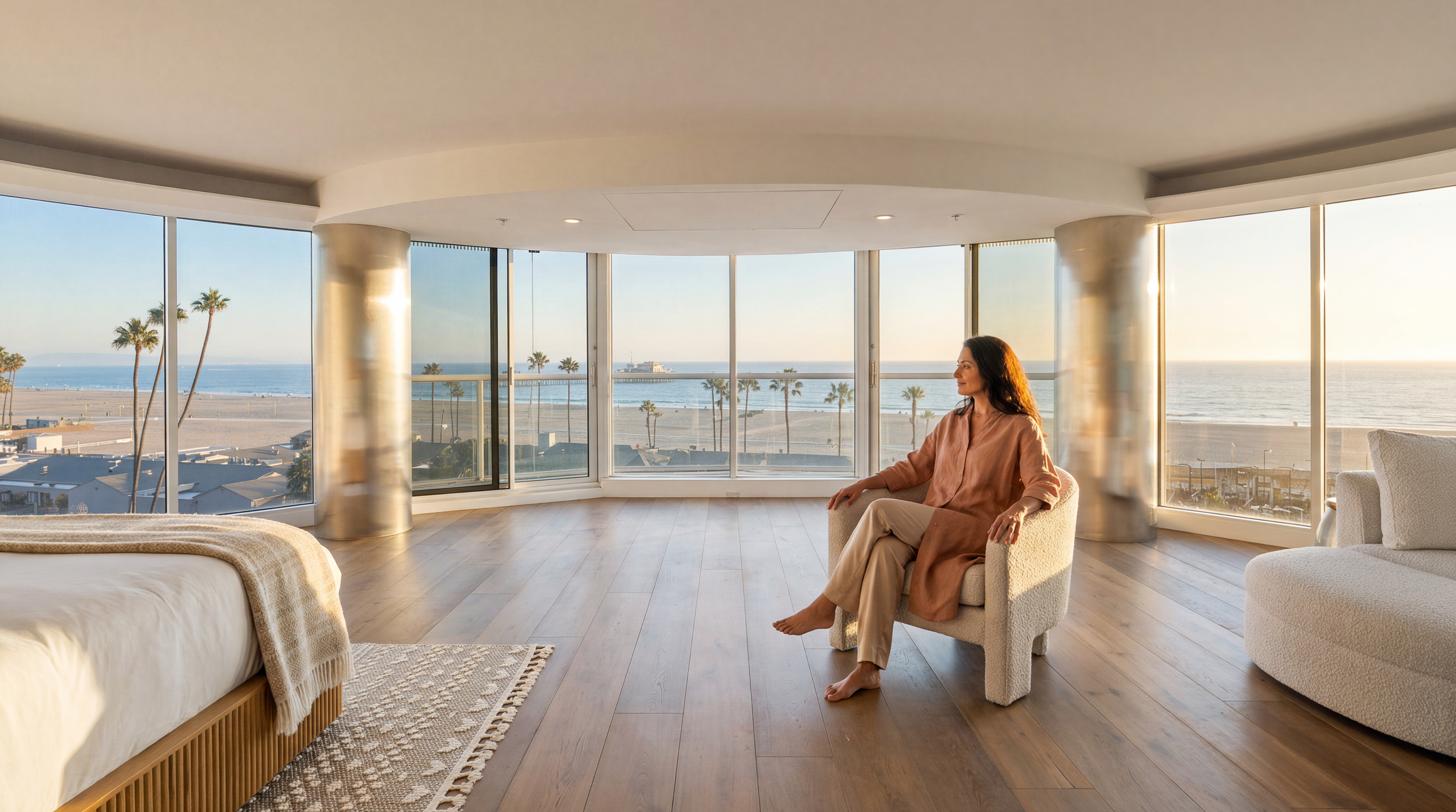 A luxurious primary bedroom featuring panoramic floor-to-ceiling windows with sweeping views of the beach and ocean. The room boasts a sophisticated, minimalist design with warm hardwood flooring, a plush armchair where a person is seated, and a neutral, coastal-inspired aesthetic. This spacious, light-filled suite emphasizes high-end indoor-outdoor living with its seamless connection to the coastal environment.