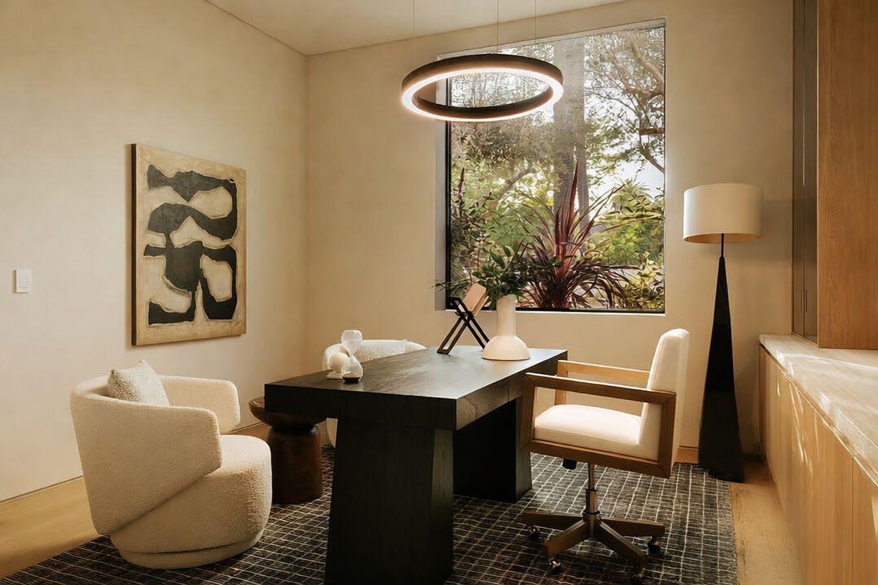 This sophisticated home office features a dark wood desk centered in the room, complemented by a cream-colored textured armchair and a modern office chair. A large, rectangular window provides a view of the outdoors, while a circular pendant light fixture hangs above the desk. The space is accented by a piece of abstract wall art and a tall floor lamp, creating a refined and professional atmosphere.