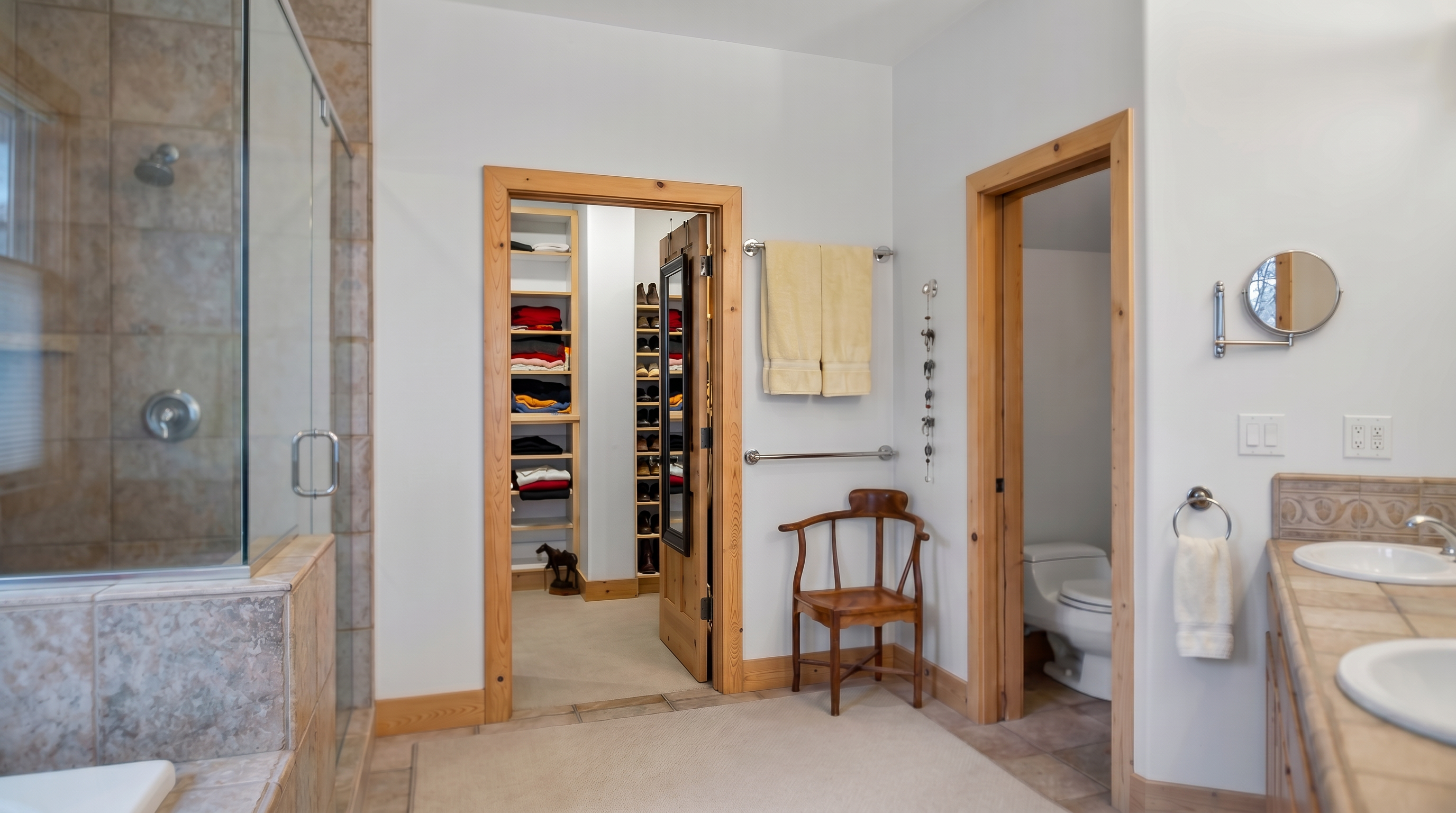 This spacious primary bathroom features natural light tones, tile flooring, and a double-sink vanity. The room includes a glass-enclosed shower, a private toilet area, and a convenient walk-in closet with built-in shelving. The wooden door frames and chair add a touch of warmth to the functional design.
