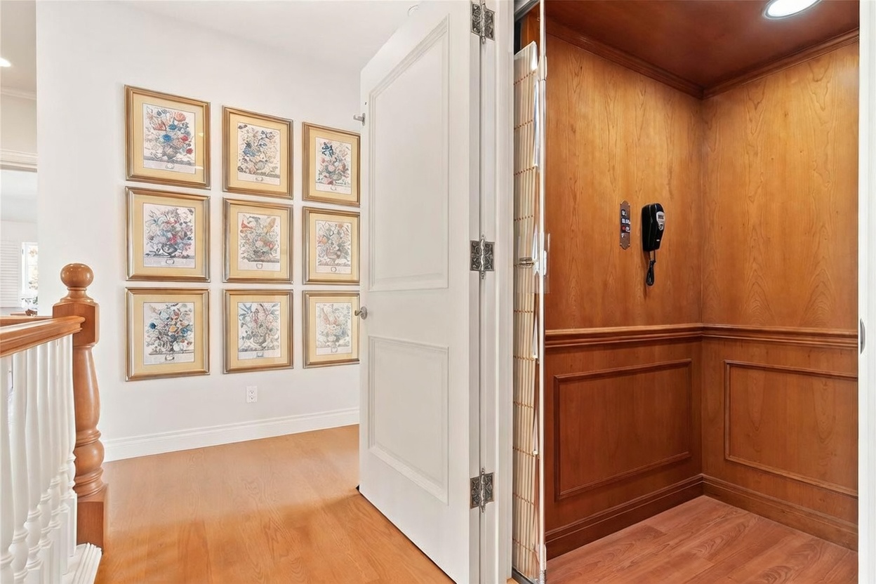 This image captures a transition space featuring a home elevator with warm wood-paneled walls and a classic phone fixture. To the left, a white door stands open, revealing a hallway wall decorated with a grid of nine framed floral prints above light wood flooring. The perspective looks from the hallway into the elevator cab, highlighting the contrast between the bright, airy corridor and the rich, traditional interior of the lift.