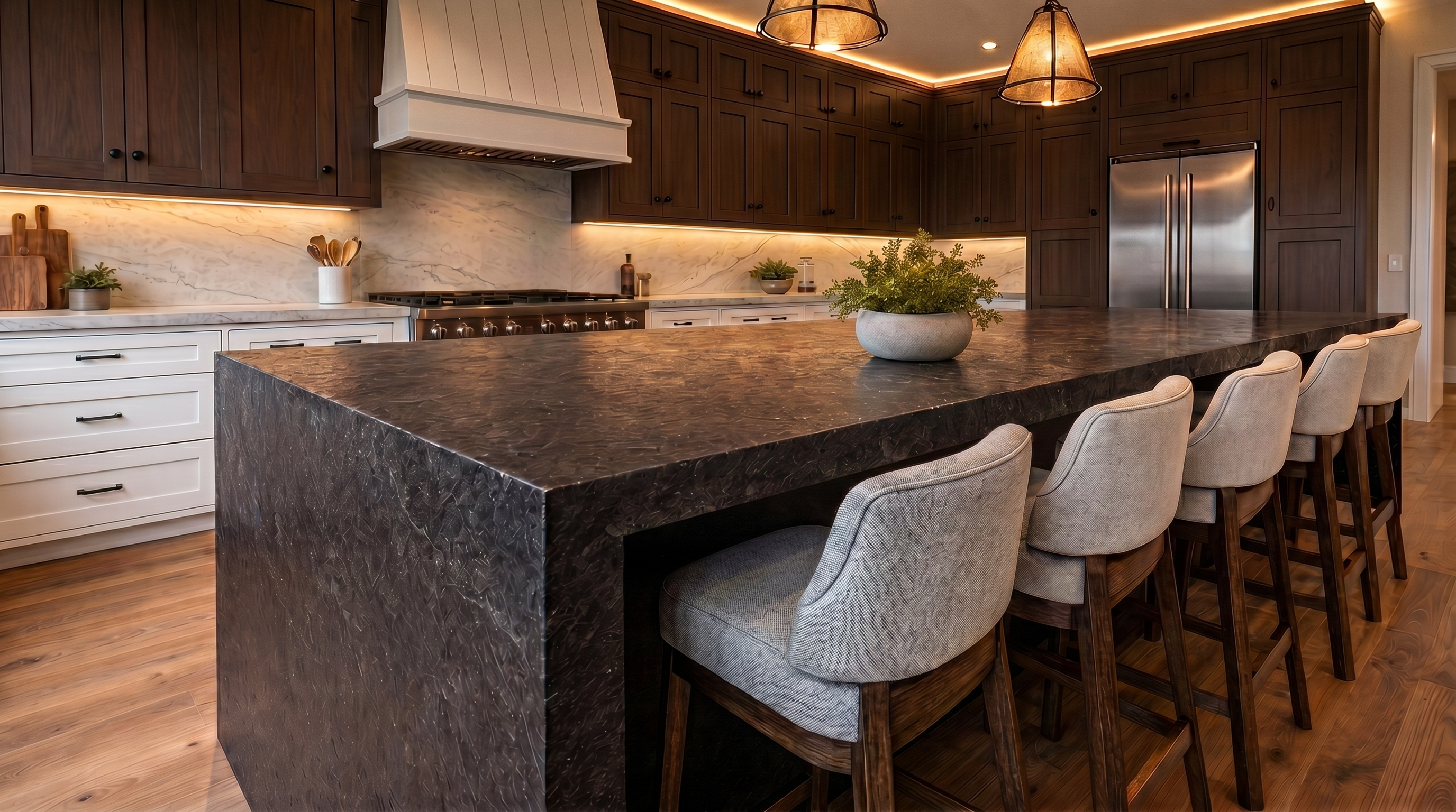 This sophisticated, modern kitchen features a spacious dark stone waterfall-edge island complemented by matching fabric bar stools. The space is anchored by warm wood-tone cabinetry, professional-grade stainless steel appliances, and elegant marble slab backsplashes with integrated under-cabinet lighting. Large, designer pendant lights highlight the center island, creating a warm and luxurious aesthetic perfect for both cooking and entertaining.