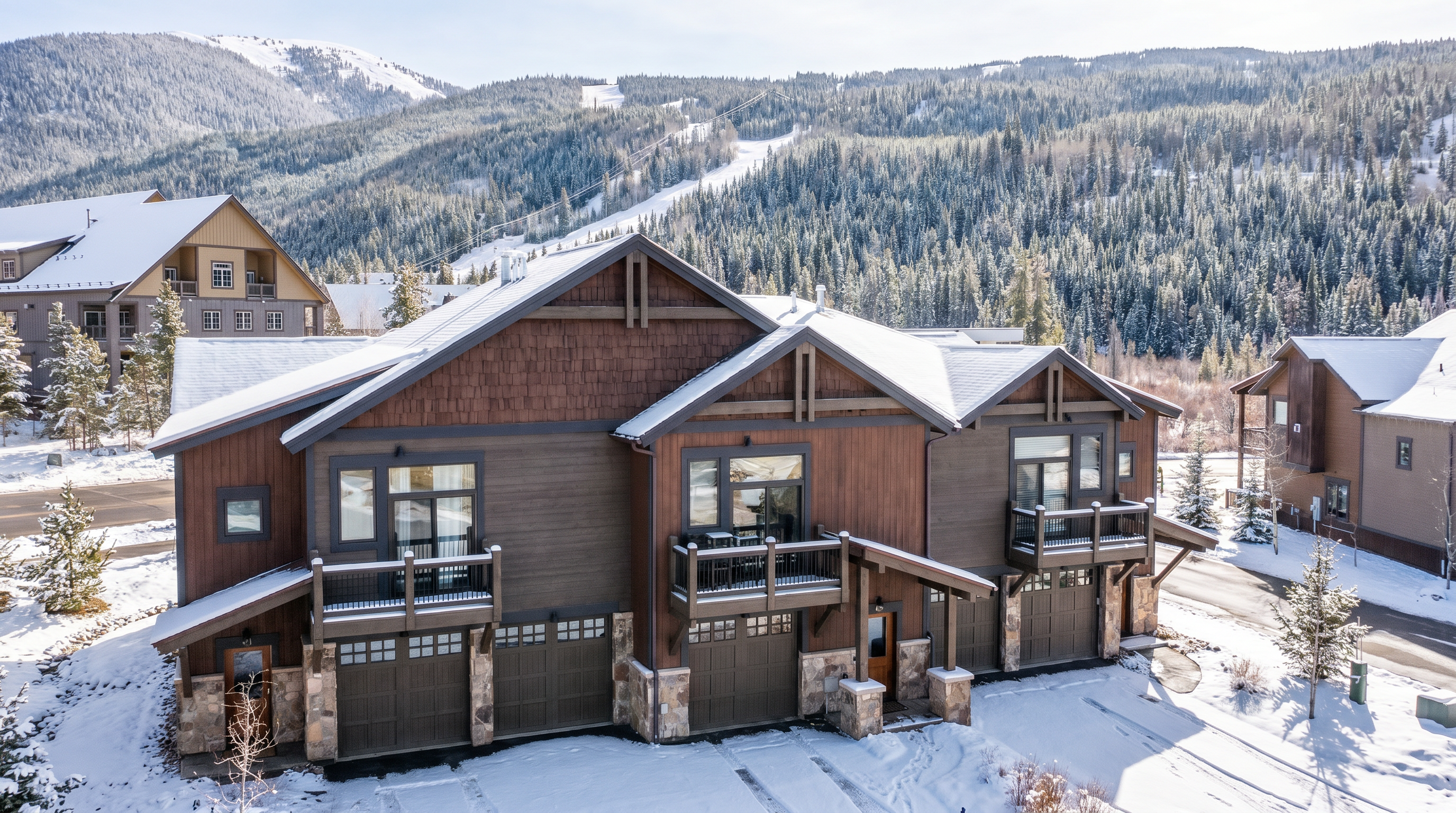 This high-angle aerial shot captures a multi-unit mountain townhome complex featuring rustic wood siding, stone accents, and dark shingled roofs. Each unit includes a private balcony and a garage, set against a backdrop of a lush pine forest and a prominent ski slope. The perspective emphasizes the property's prime location in a scenic, mountainous resort environment.