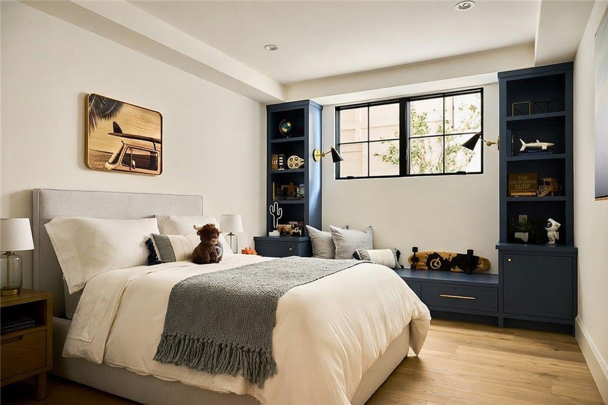 This cozy bedroom features a neutral color palette with a light-toned upholstered bed and soft bedding. Built-in navy blue shelving units flank a window, providing both storage and a decorative focal point, while light wood flooring adds warmth to the space. The room is accented with playful decor, including a surfboard-themed wall art piece and a skateboard, creating a relaxed and inviting atmosphere.