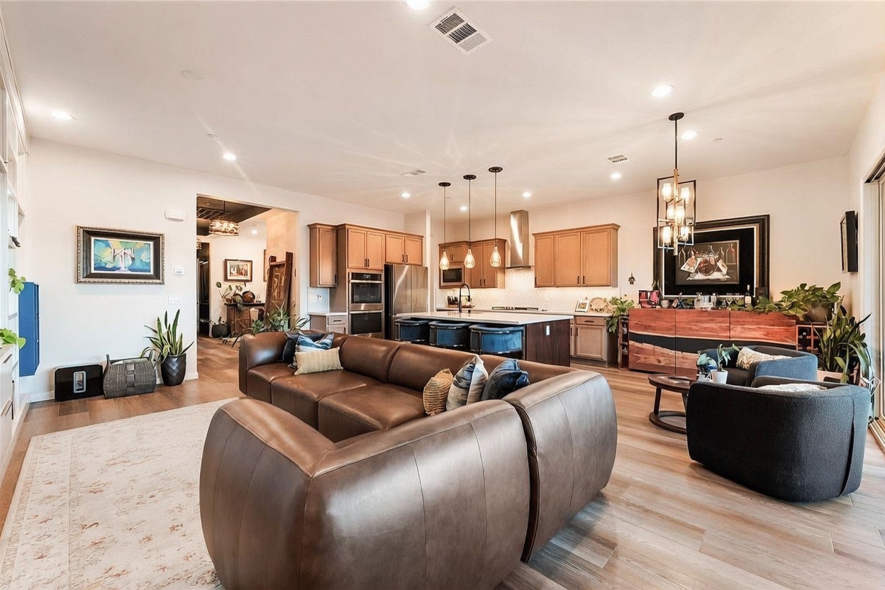 This open-concept living area features a large, plush brown leather sectional sofa as the central focal point, positioned on a light-toned area rug. The space flows seamlessly into a kitchen equipped with light wood cabinetry, a central island with bar seating, and modern pendant lighting. The room is decorated with contemporary art, indoor plants, and a wooden sideboard, creating a warm and inviting atmosphere with a clear view into the adjacent living spaces.