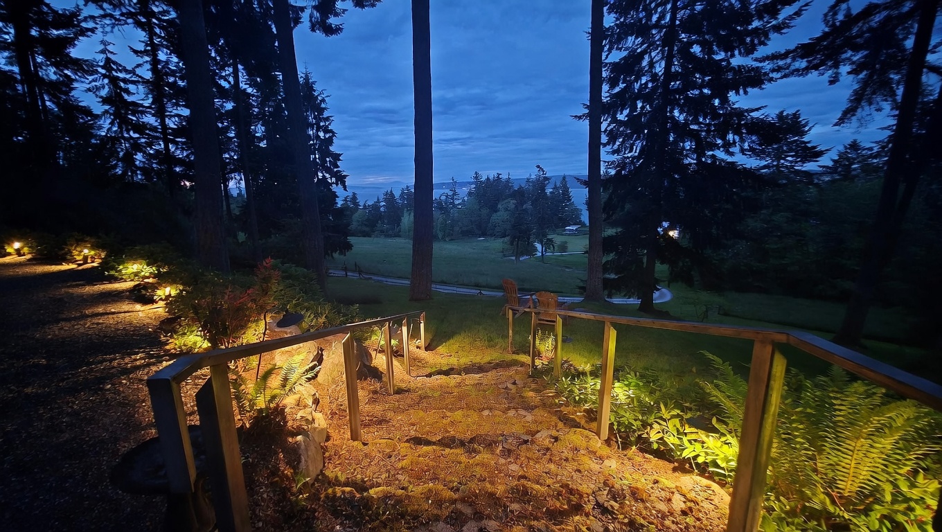 This twilight view captures a set of stone steps leading down from a landscaped terrace into a sprawling, grassy backyard framed by tall evergreen trees. The scene is illuminated by warm, low-voltage landscape lighting that highlights the path and surrounding foliage, creating an inviting and serene atmosphere. In the distance, the silhouette of the landscape opens up to reveal a glimpse of the water under a dusky blue sky.