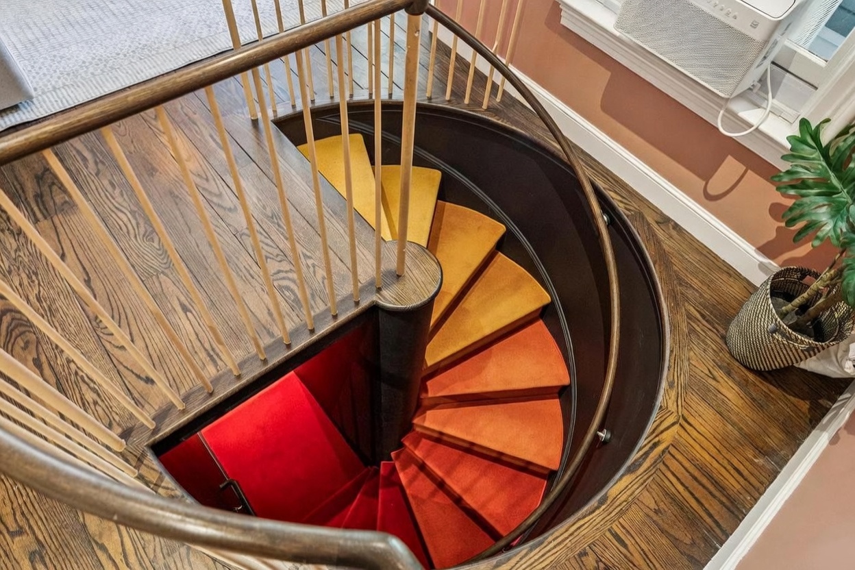 This high-angle shot captures a striking spiral staircase that serves as a bold architectural focal point. The steps feature a vibrant, gradient carpet runner that transitions from deep red at the bottom to bright yellow at the top, framed by dark wood treads and a curved wooden handrail. The surrounding floor is finished in warm-toned wood planks, and a potted plant sits nearby, adding a touch of greenery to the space.