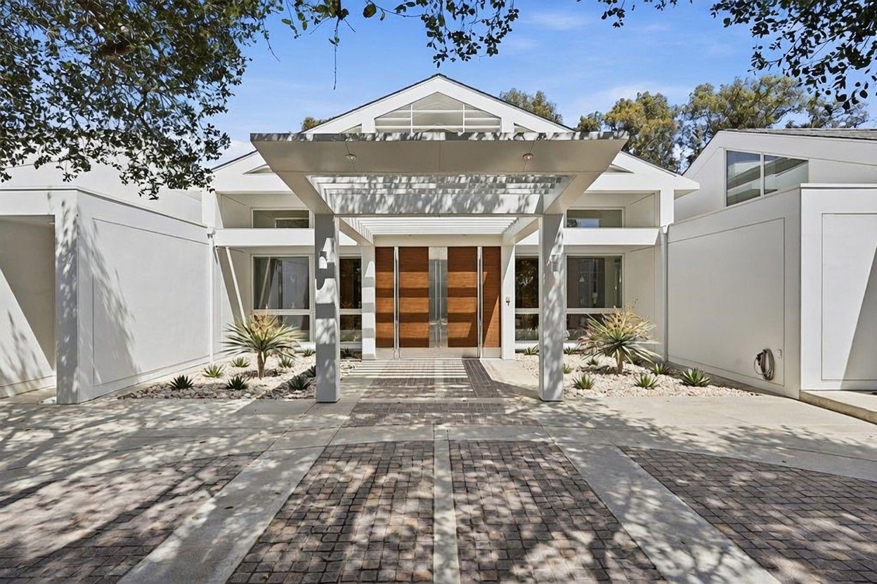 This striking modern home features a prominent, covered entryway defined by a geometric, white-framed portico that leads to a set of double wooden doors. The front facade is characterized by clean, white lines, large windows, and a symmetrical design, complemented by a paved driveway and landscaped garden beds with desert-style plants. The perspective is a straight-on, eye-level shot that emphasizes the architectural symmetry and the inviting, upscale nature of the entrance.