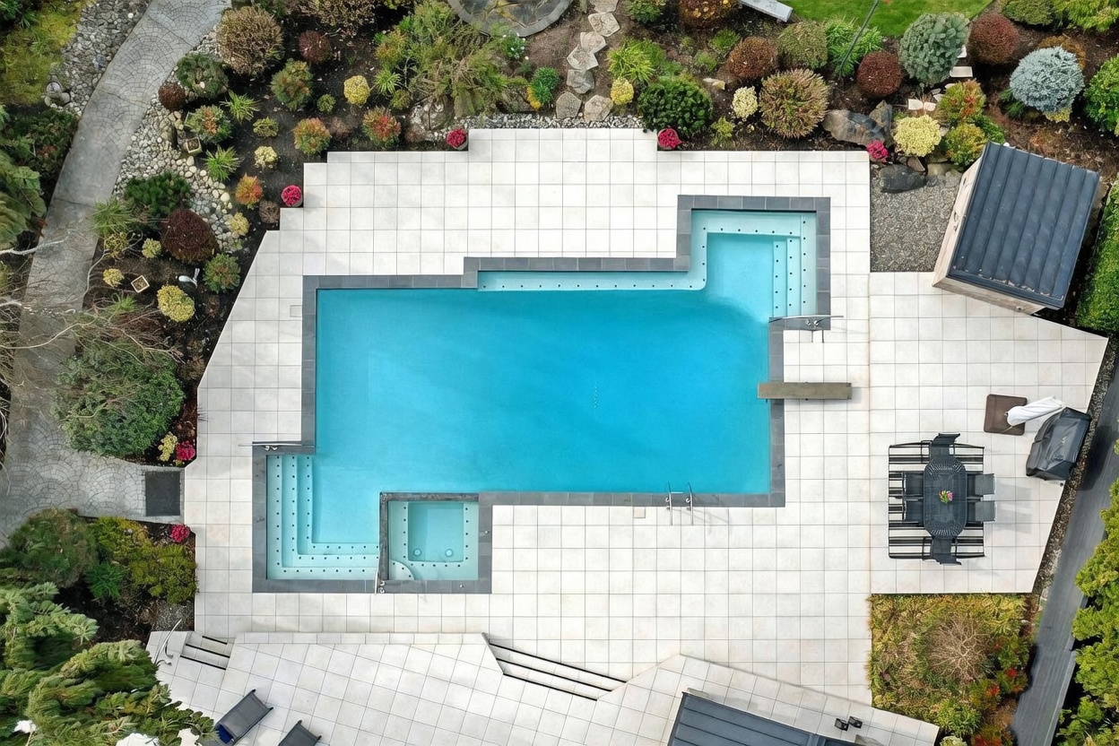 This high-angle aerial view captures a luxurious backyard swimming pool and spa complex surrounded by pristine light-colored stone tiling. The pool features a unique geometric design with integrated steps and a diving board, complemented by lush, manicured landscaping and a small outdoor dining area. The perspective provides a clear, comprehensive look at the outdoor entertainment space, emphasizing its clean lines and resort-like atmosphere.