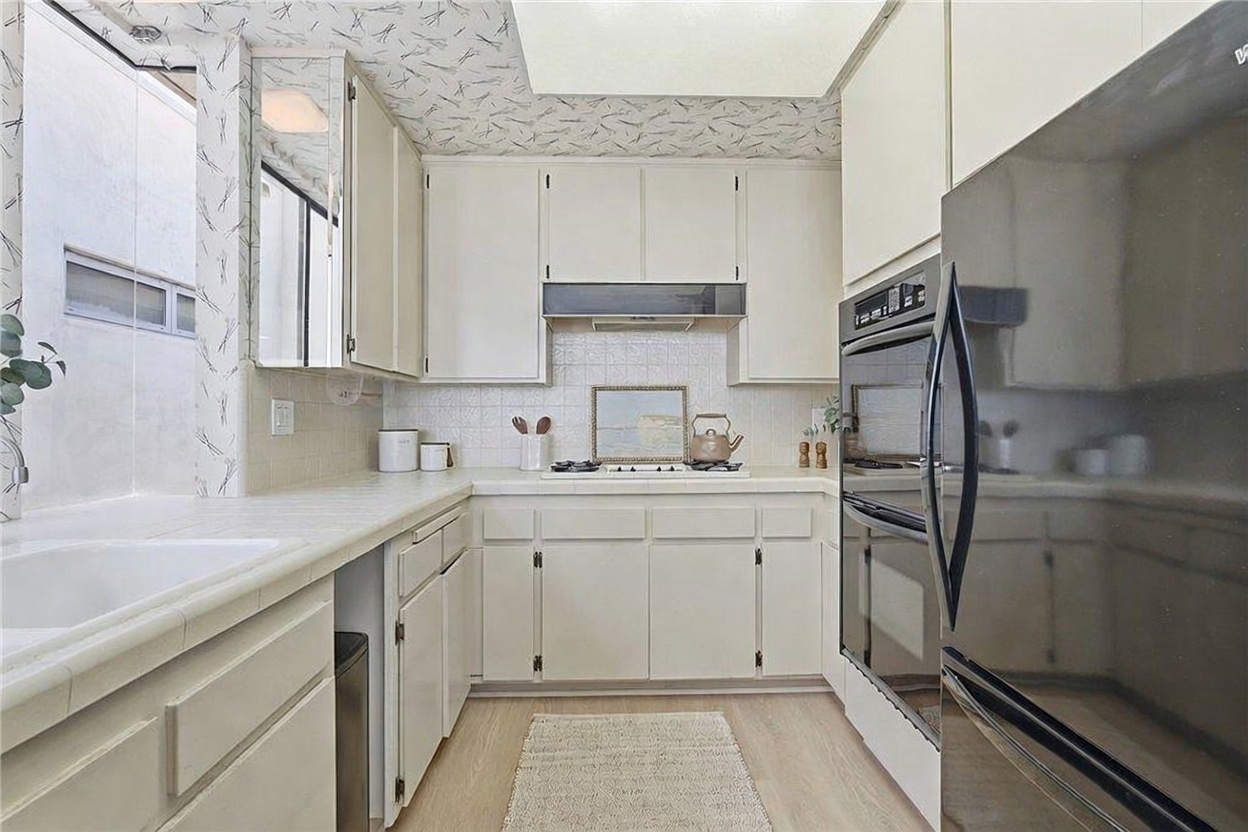This U-shaped kitchen features light-colored cabinetry, a tiled backsplash, and a built-in double wall oven. The space includes a light-toned countertop, a cooktop, and a neutral-toned floor, creating a bright and functional workspace. The perspective is a straight-on view from the kitchen entrance, highlighting the efficient layout and classic design.