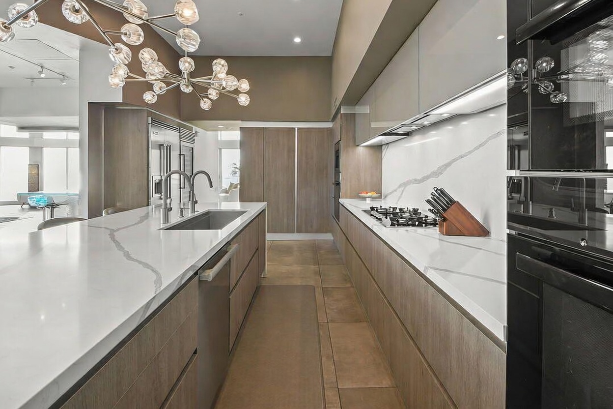 This modern, high-end kitchen features a long central island with a stone countertop and an integrated sink, complemented by light wood cabinetry and sleek, dark-toned appliances. A striking, multi-bulb chandelier hangs above the island, while a gas cooktop is set into the stone counter along the back wall. The perspective is a straight-on view looking down the length of the kitchen, highlighting the clean lines and contemporary design.
