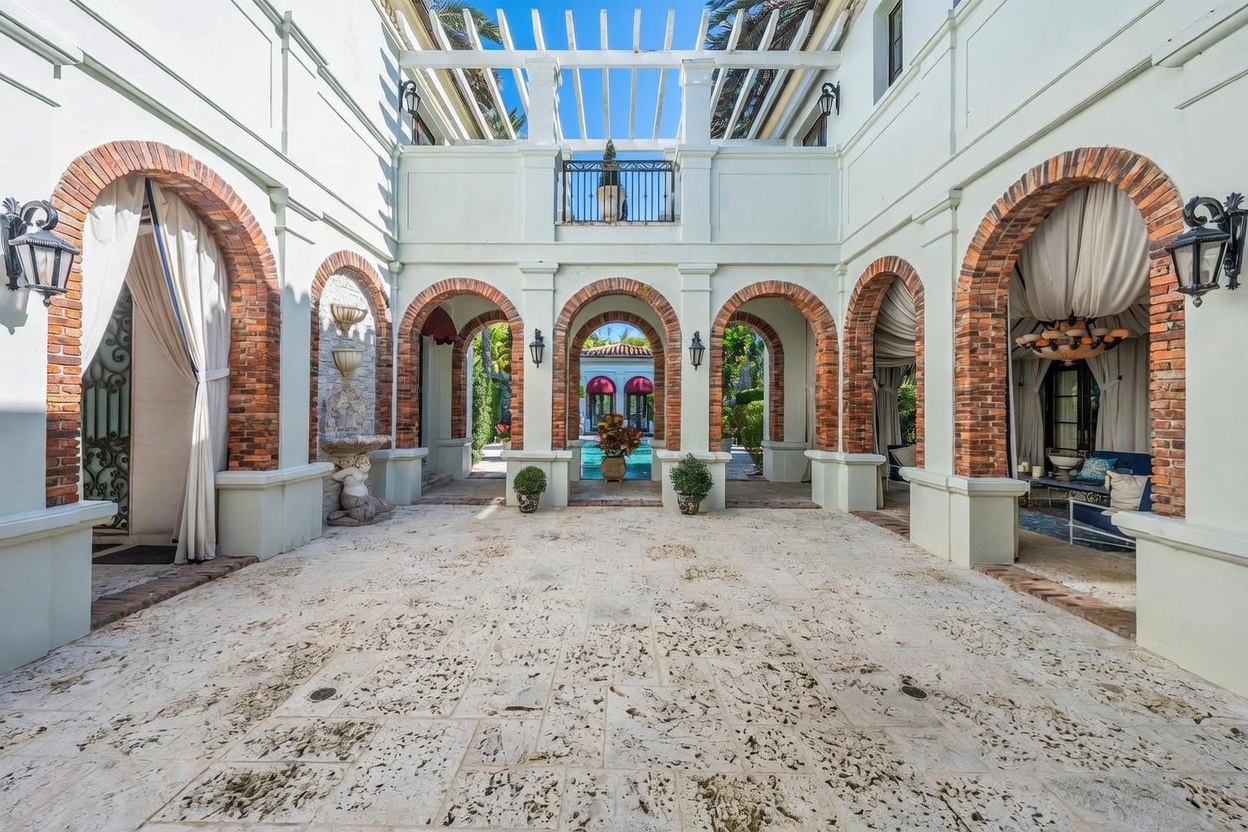 This elegant Mediterranean-style courtyard features a central stone-paved patio framed by a series of brick-accented arches. A pergola structure spans the upper level, while a tiered fountain and lush potted plants add a sophisticated touch to the outdoor living space. The perspective looks through the arched colonnade toward a swimming pool area, creating a seamless transition between the covered patio and the open-air backyard.