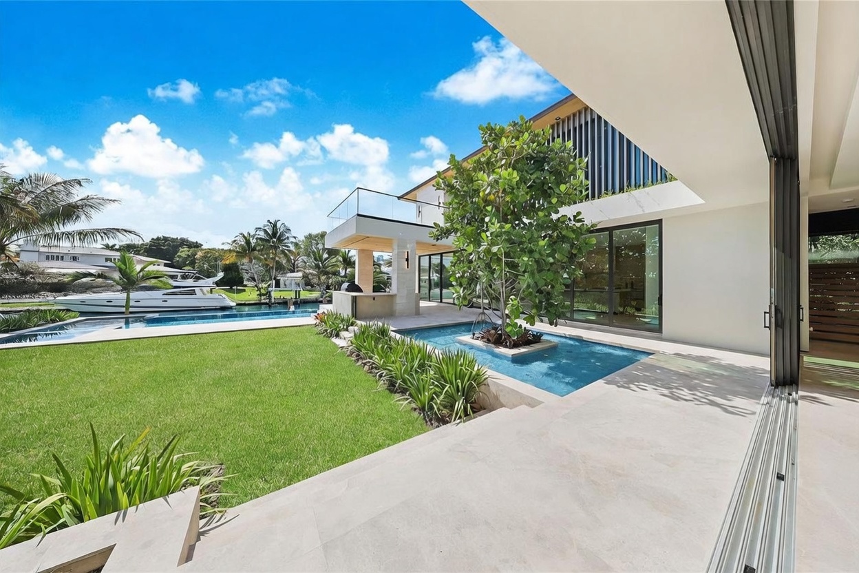 This modern waterfront estate features a seamless transition between indoor and outdoor living spaces, highlighted by large sliding glass doors that open onto a stone patio. The backyard includes a manicured lawn, a custom water feature with a central tree, and a swimming pool that overlooks a canal with a docked yacht. The architectural design emphasizes clean lines, expansive glass, and a sophisticated, resort-like atmosphere.
