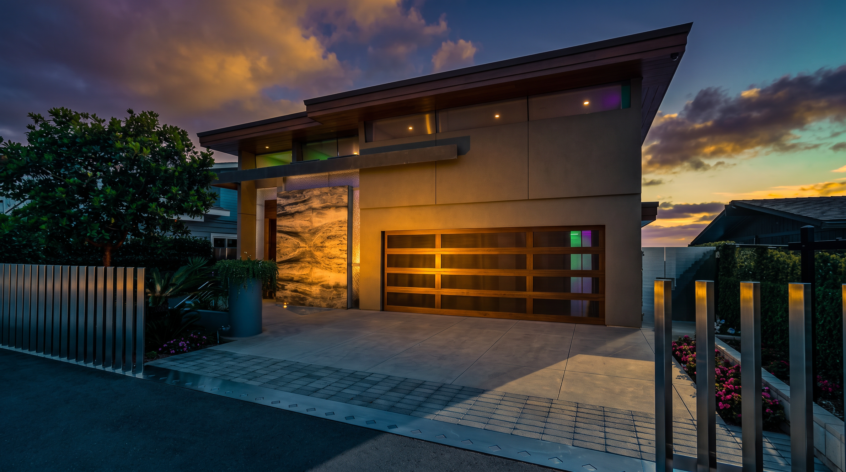 This modern residence features a striking architectural design with a prominent stone-clad accent wall near the entrance and a sleek, multi-panel garage door. The exterior is finished in a neutral, smooth-textured material, complemented by warm wood-toned soffits and horizontal clerestory windows. A contemporary metal fence frames the foreground, leading to a clean concrete driveway and a landscaped garden area with vibrant red flowers.