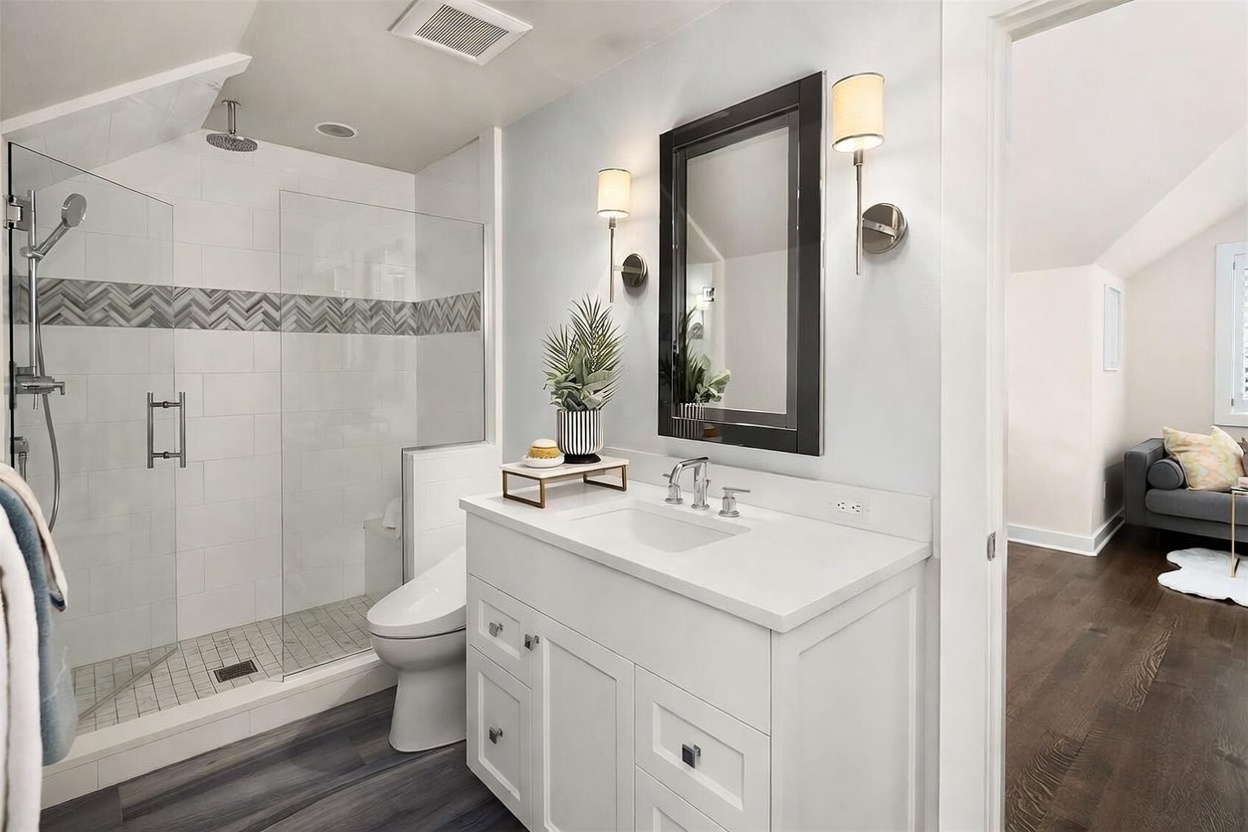 This bright, modern bathroom features a white vanity with a stone countertop and a sleek, rectangular mirror flanked by two wall-mounted light fixtures. To the left, a glass-enclosed walk-in shower showcases white subway tile walls with a decorative chevron-patterned accent band and a rainfall showerhead. The room is finished with dark wood-look flooring, creating a clean and contemporary aesthetic that flows into an adjacent living space.