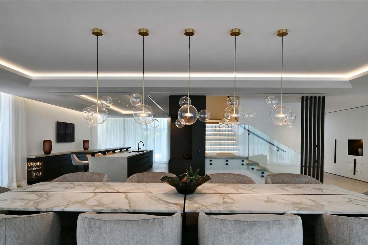 This sophisticated dining area features a long, light-toned stone table surrounded by plush, upholstered chairs, set beneath a series of modern, multi-globe pendant lights. The open-concept layout flows seamlessly into a sleek kitchen with a large island and a striking staircase with glass railings in the background. The space is defined by a clean, contemporary aesthetic with neutral tones and recessed lighting, creating an elegant and airy atmosphere.