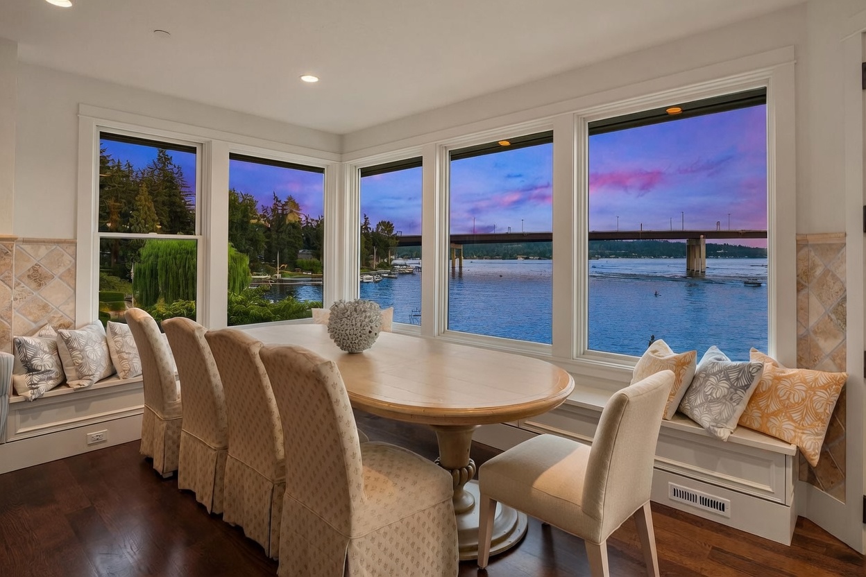 This elegant dining area features a large oval wooden table surrounded by six upholstered chairs, positioned to take full advantage of the expansive water views. Large windows wrap around the space, showcasing a scenic sunset over the lake and a bridge in the distance. Built-in window seats with patterned cushions provide additional seating, creating a cozy and inviting atmosphere for waterfront dining.