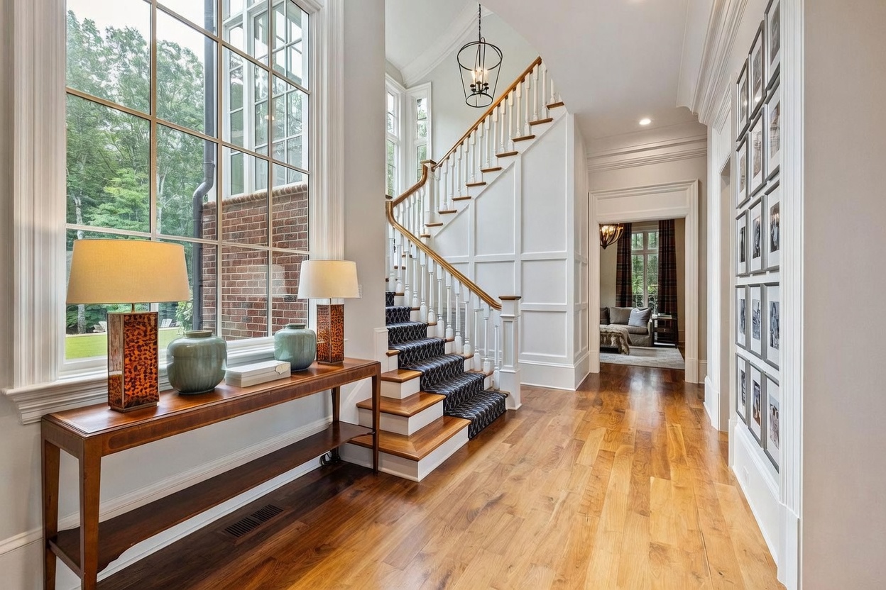 This elegant foyer features a grand staircase with a patterned runner and a wooden handrail, serving as the focal point of the entryway. To the left, a console table with matching lamps sits beneath large windows that provide ample natural light, while a gallery wall of framed photos lines the hallway on the right. The space is finished with light wood flooring and crisp white trim, leading the eye toward a secondary room visible in the background.