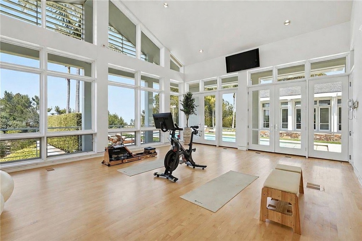 This bright and airy home gym features light wood flooring and an expansive wall of glass that provides abundant natural light and views of the outdoors. The space is equipped with a stationary exercise bike and a rowing machine, with a wooden bench positioned in the foreground. The high, vaulted ceiling and multiple glass doors create an open, inviting atmosphere suitable for a workout area.