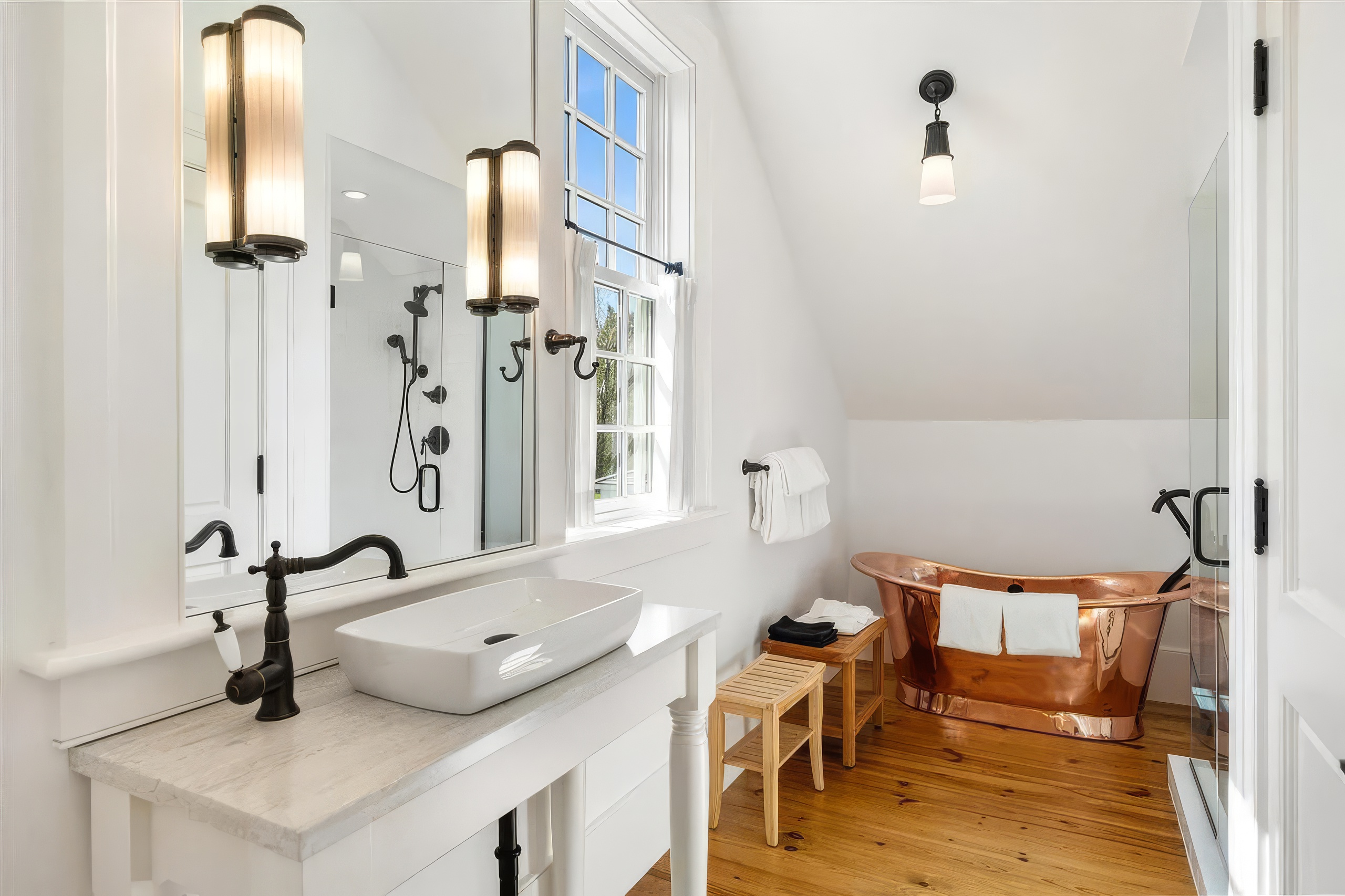 This bright and airy bathroom features a striking copper soaking tub as its centerpiece, set against a backdrop of warm wood flooring and white walls. A sleek vessel sink sits atop a stone-topped vanity, complemented by dark metal fixtures and wall-mounted lighting. The space is illuminated by natural light from a window, creating a clean, sophisticated, and inviting atmosphere.