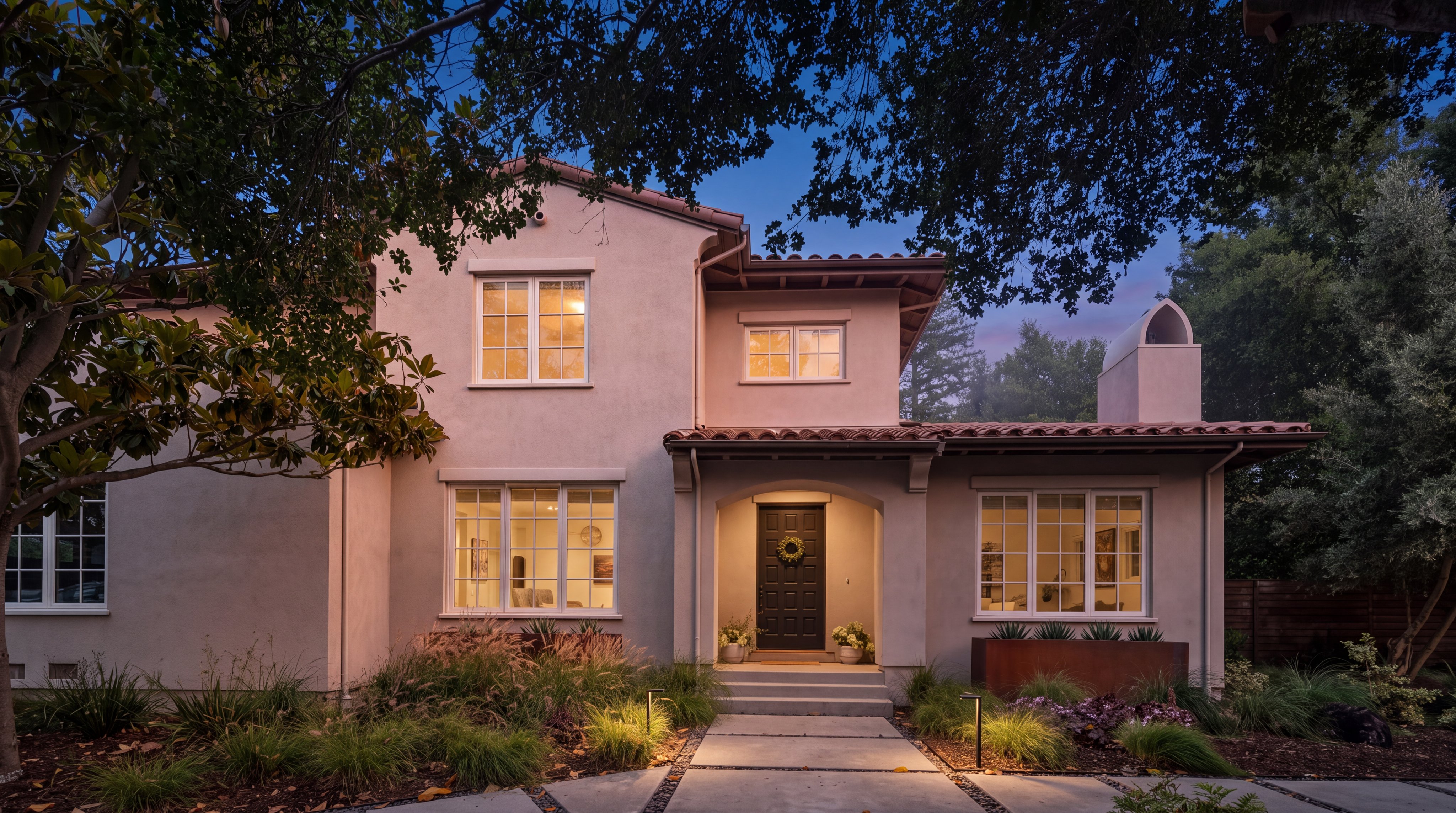 This two-story home features a light-colored stucco facade with a classic Mediterranean-inspired design, characterized by a terracotta tile roof and a symmetrical layout. A concrete walkway leads through a landscaped front yard with native grasses and metal planters to a dark-toned front door set within an arched entryway. The scene is captured from a ground-level perspective, framed by the dappled shade of mature trees that cast soft shadows across the front of the house.
