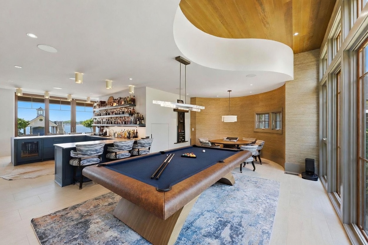 This expansive entertainment space features a prominent pool table as the central focal point, set atop a large patterned area rug. To the left, a well-stocked wet bar with dark cabinetry and bar seating overlooks a scenic waterfront view through large windows. The room is defined by a unique curved ceiling design with warm wood paneling and a textured, curved wall that creates a sophisticated, lounge-like atmosphere.