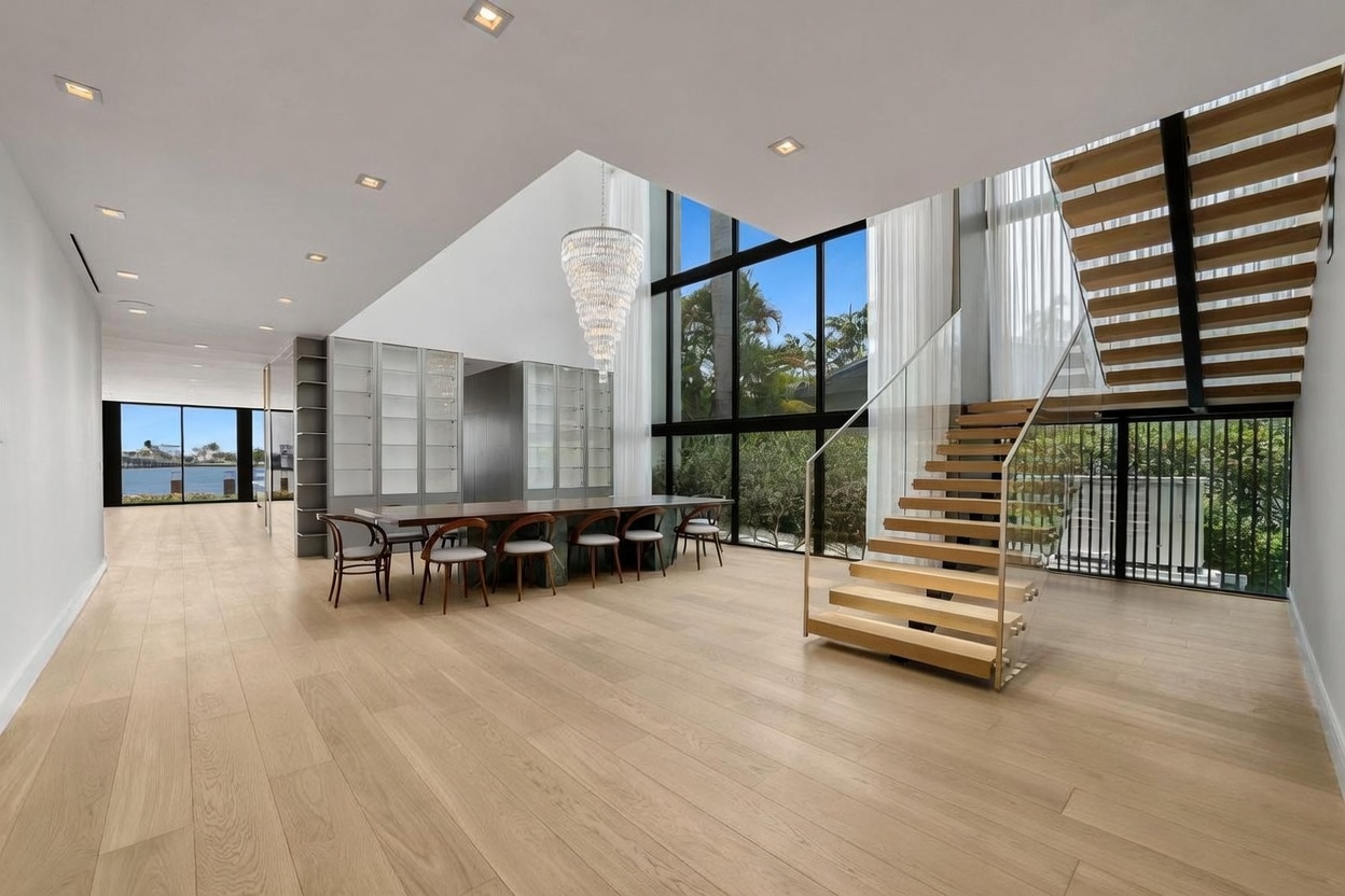 This expansive, open-concept dining area features light wood flooring and a dramatic, double-height ceiling anchored by a large, multi-tiered crystal chandelier. A long dining table with six chairs sits adjacent to a wall of floor-to-ceiling windows that provide abundant natural light and views of the lush greenery outside. To the right, a modern floating staircase with glass railings adds a sculptural element to the bright, airy space.
