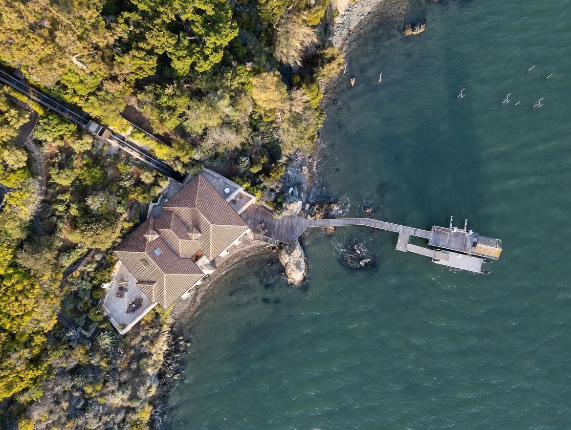 This high-angle aerial view captures a waterfront estate nestled along a lush, tree-lined coastline. The property features a multi-gabled residence with a terracotta-tiled roof, a private wooden pier extending into the water, and a funicular track descending through the dense foliage. The scene emphasizes the secluded, tranquil nature of the location and its direct access to the bay.
