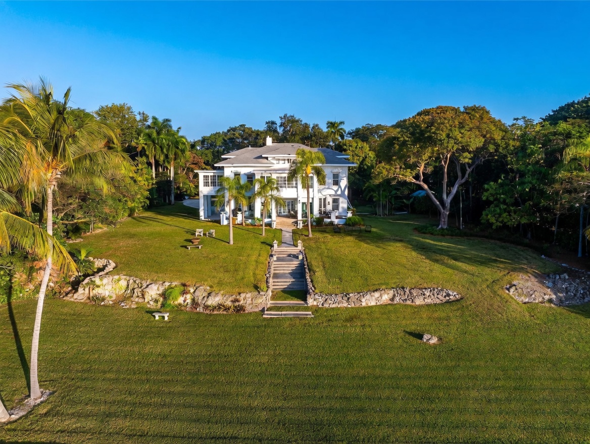 This aerial perspective captures a grand, white colonial-style estate set against a lush, expansive lawn and dense surrounding foliage. A stone-walled terrace with a central staircase leads from the lower lawn up to the main residence, which features a symmetrical facade, multiple columns, and a classic hip roof. The scene is bathed in warm, golden-hour sunlight, highlighting the manicured grounds and the stately architectural presence of the home.